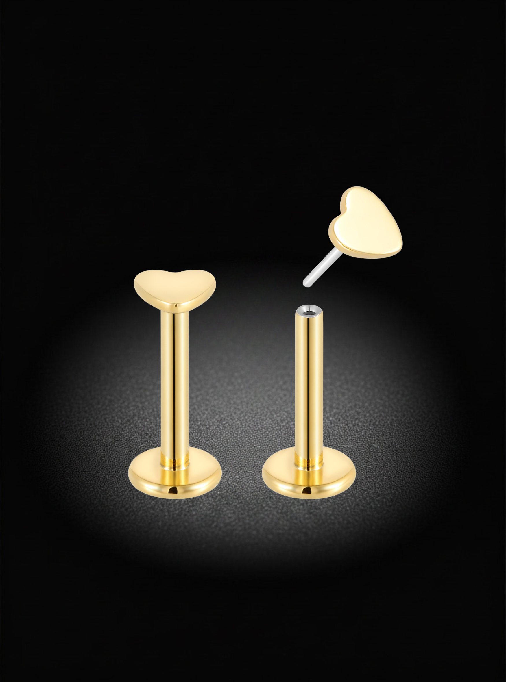 Gold heart-shaped labret ear piercings shown on a black background, with one fully assembled and one partially disassembled to show the post and flat heart tip.