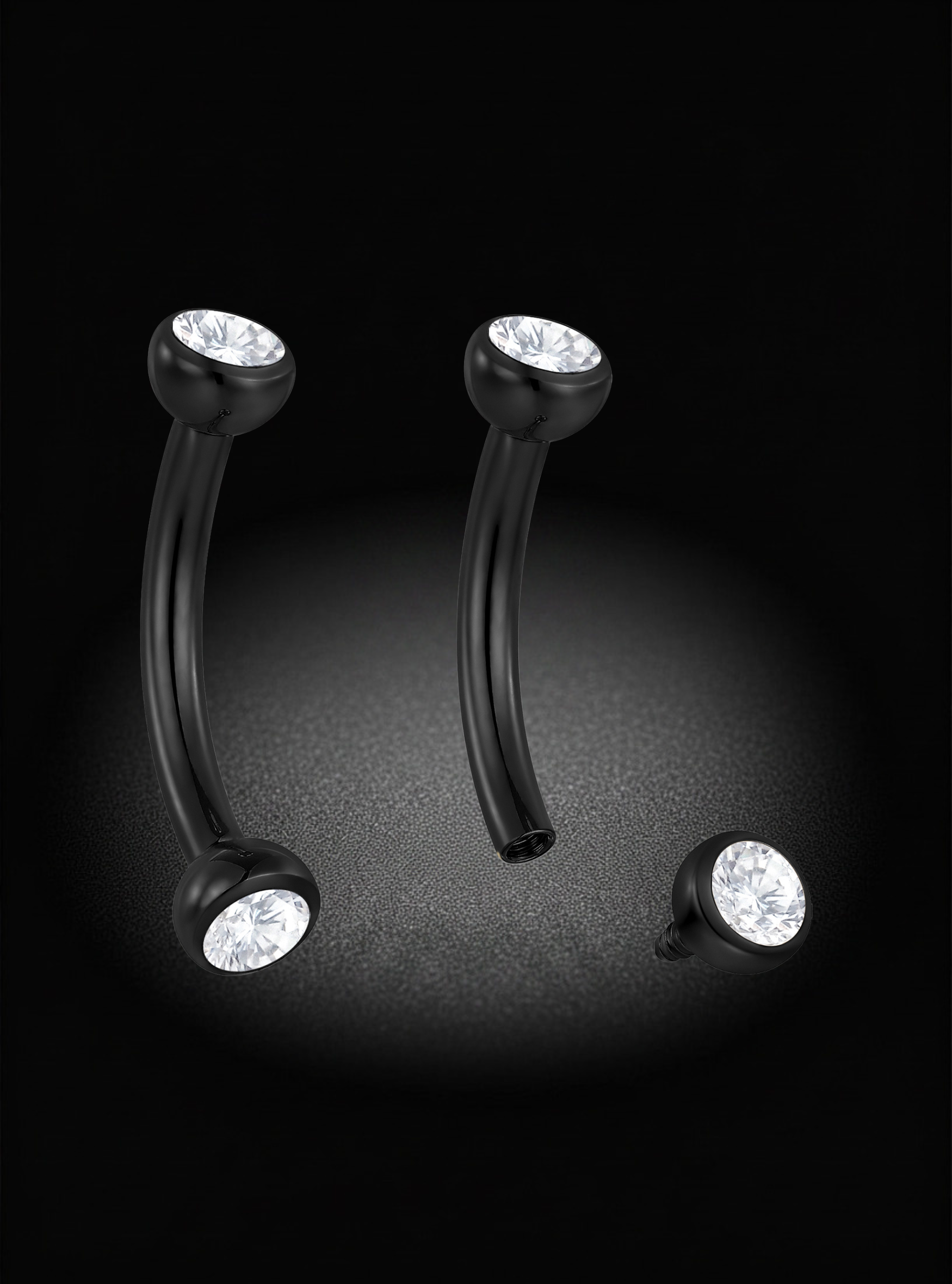 Two curved black barbell-style body jewelry pieces with clear faceted gemstones at the ends, shown on a black background with a soft spotlight effect.