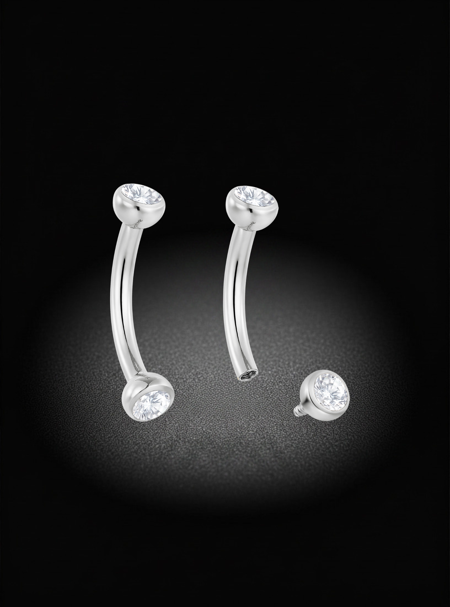 A pair of curved barbell-style body jewelry with clear gemstone end caps, shown on a black background with one piece separated to display the post.