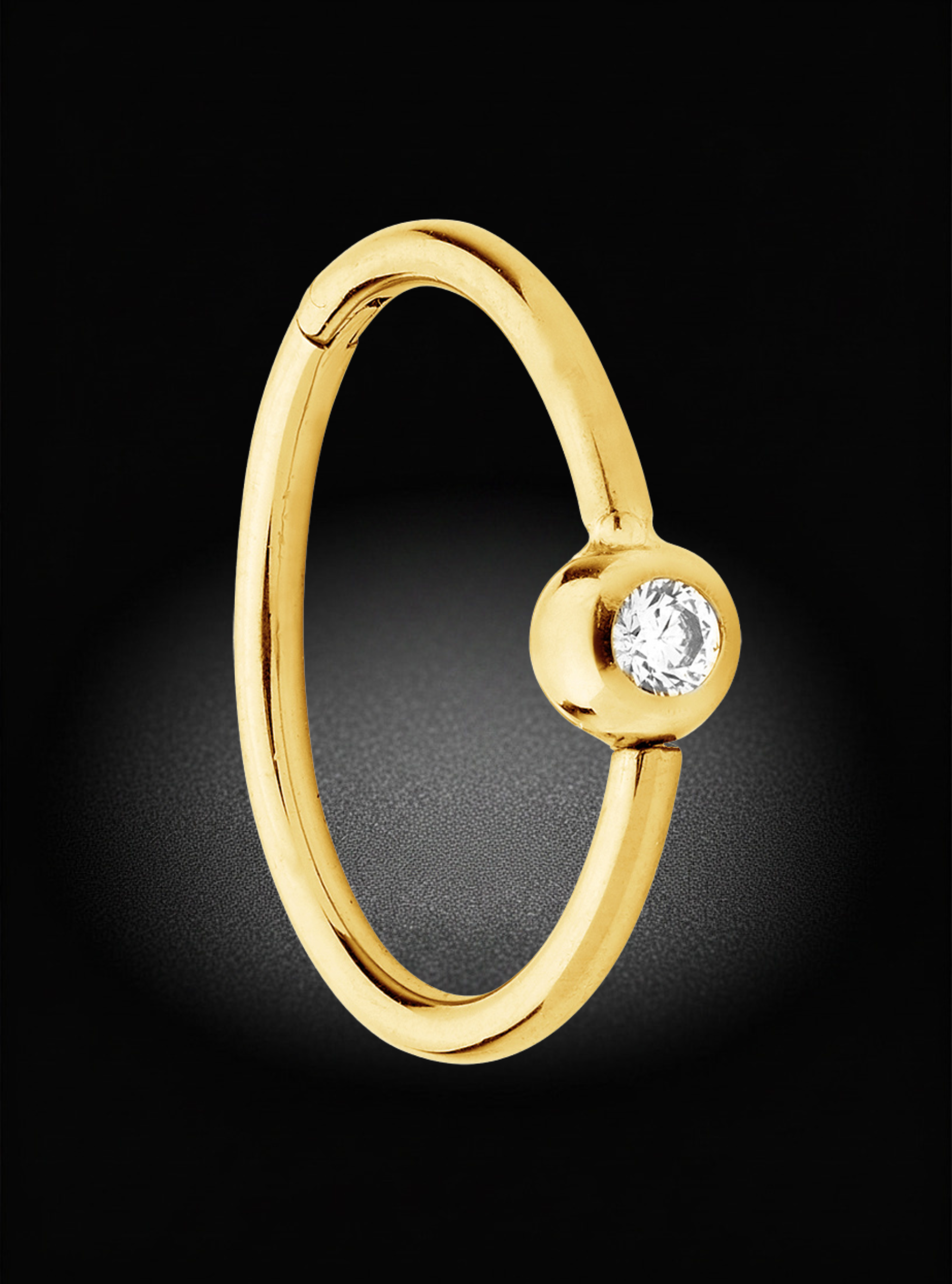A polished yellow gold huggie ring with a single bezel-set round clear gemstone, shown against a dark black background with a soft spotlight effect.