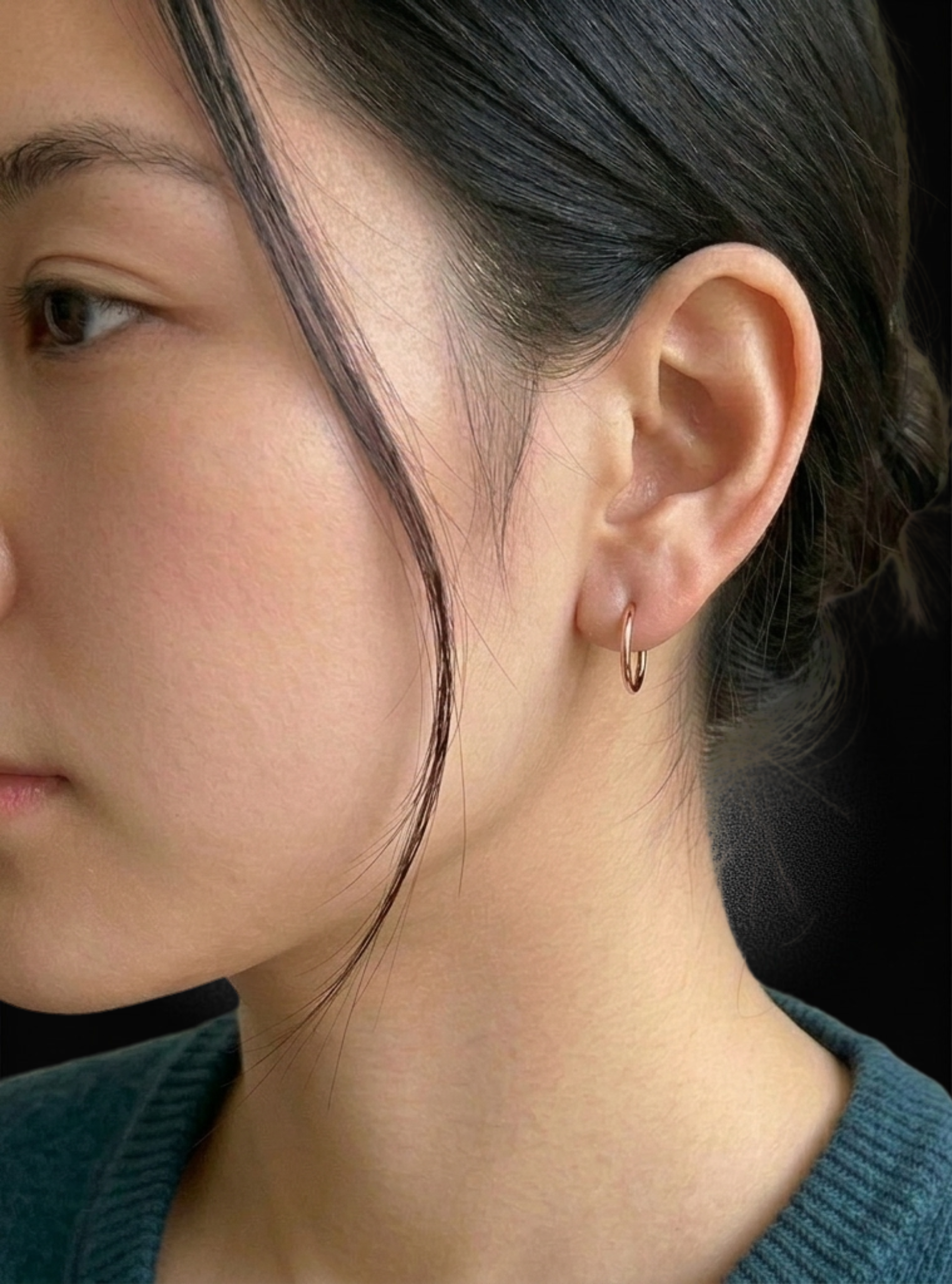 Close-up of a woman's ear wearing a small gold hoop earring, with her dark hair pulled back and a dark green ribbed top visible against a dark background.