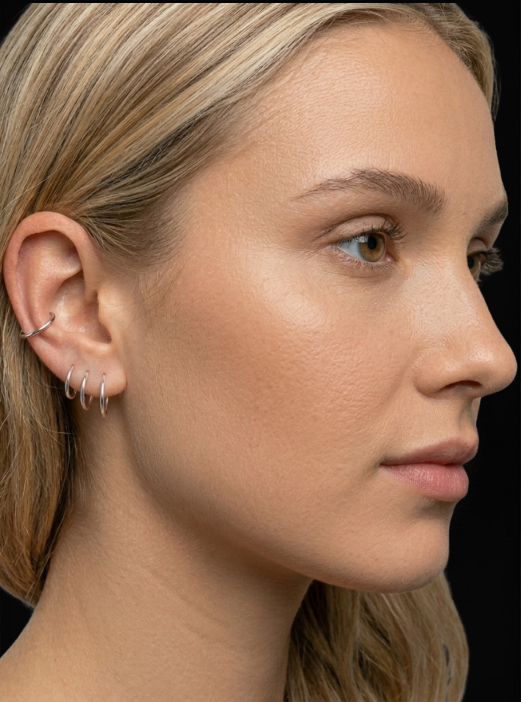 Close-up side profile of a woman with multiple silver hoop and curved ear piercings on her ear, shown against a dark background.