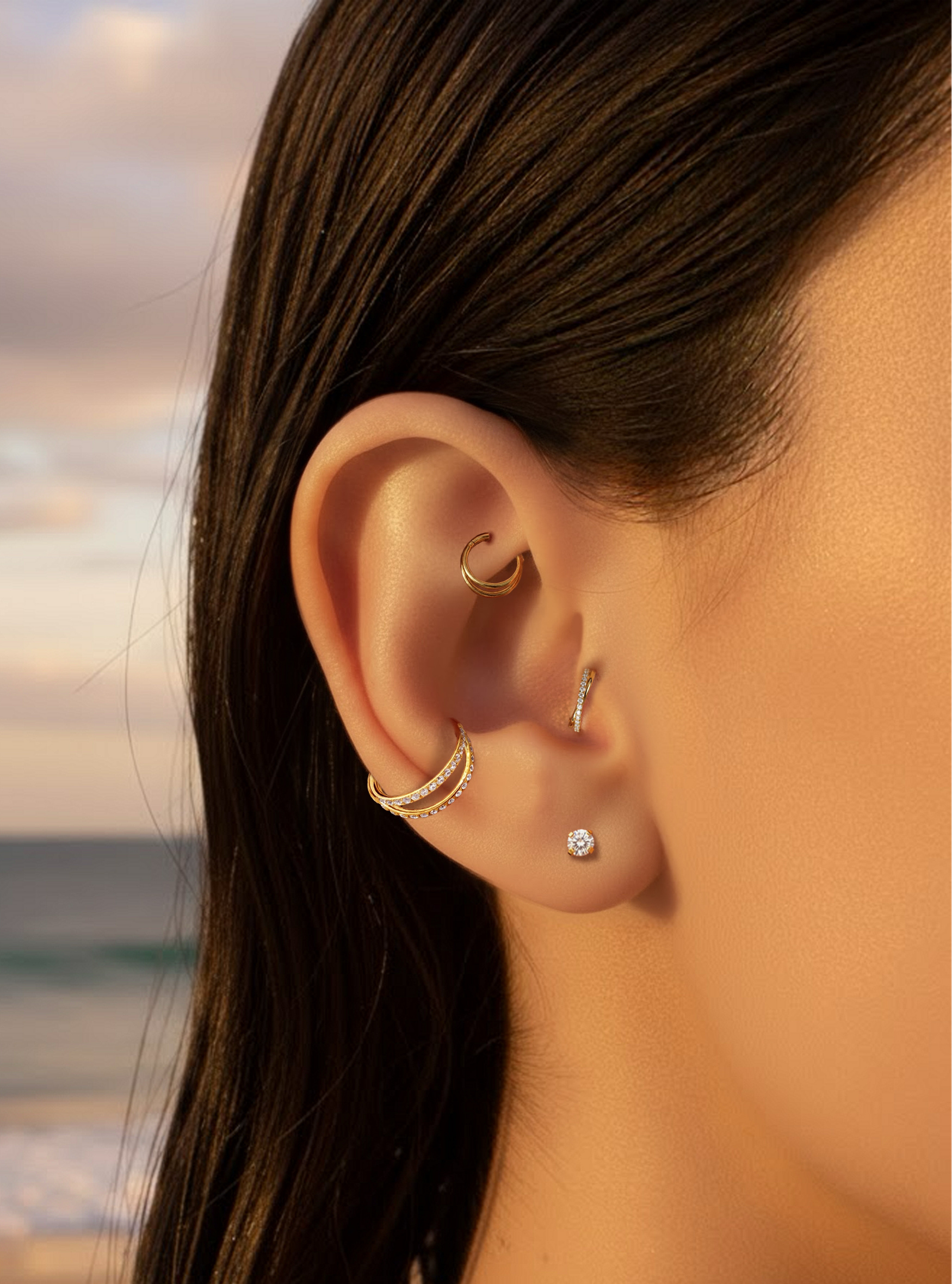 A close-up of a person's ear adorned with multiple delicate gold and silver ear piercings, including hoop, helix, lobe, and ear cuff styles, set against a softly blurred beach sunset background.