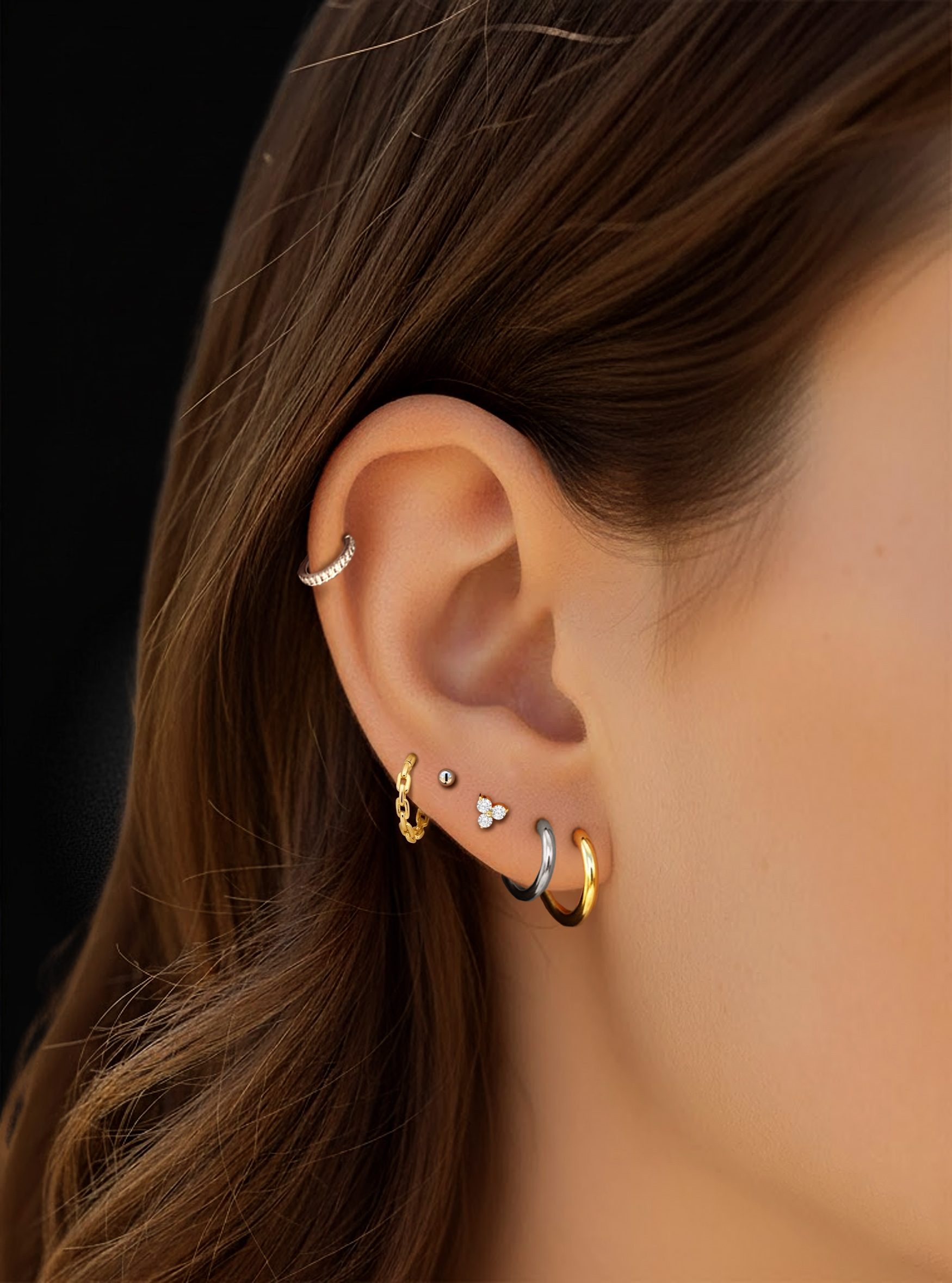 Close-up of a person's ear showcasing multiple gold and silver ear piercings, including hoop, cartilage, and lobe studs, against dark brown hair and a black background.