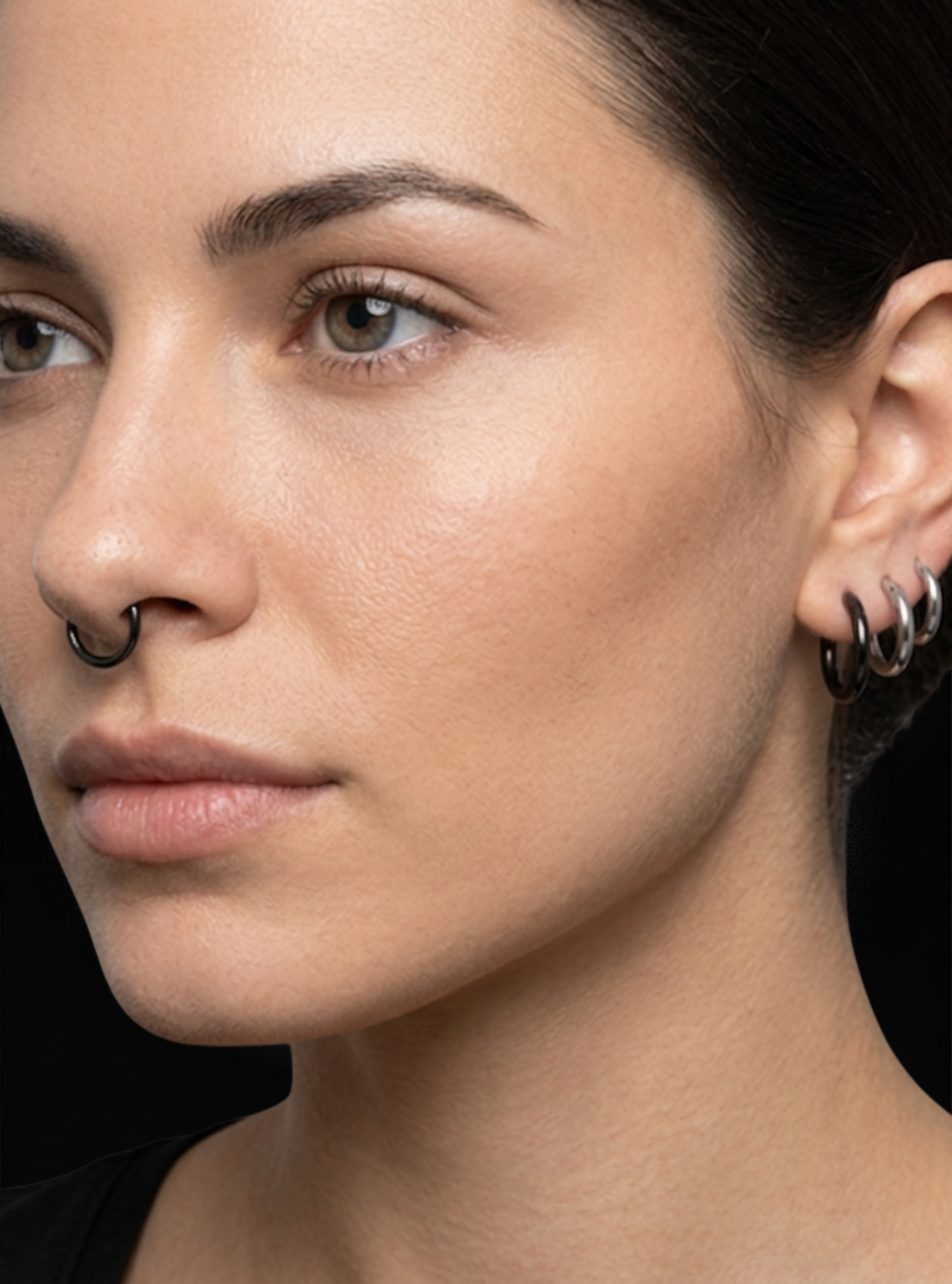 Close-up of a woman's face and ear showing black metal piercings, including a nose ring and stacked hoop earrings.