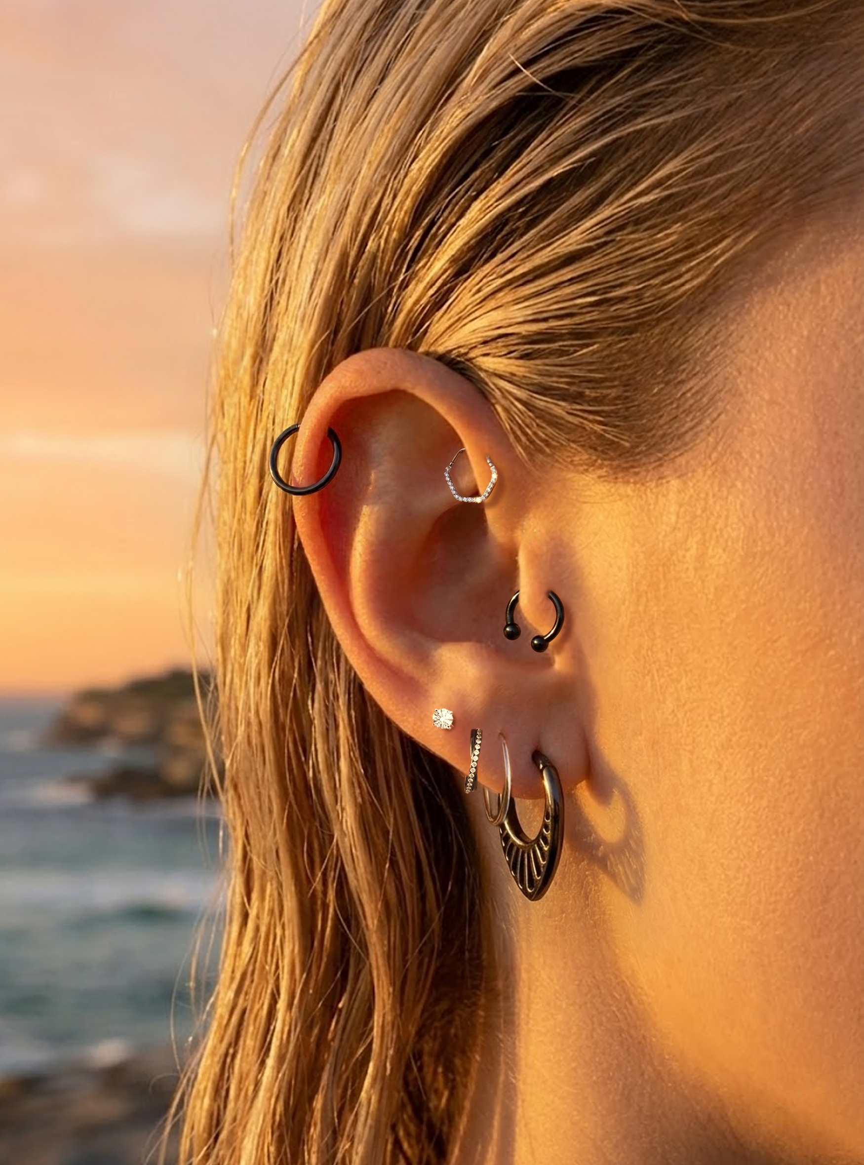 Close-up of a person's ear wearing multiple piercings and hoop earrings, including black and silver pieces, with a beach and ocean in the background at sunset.