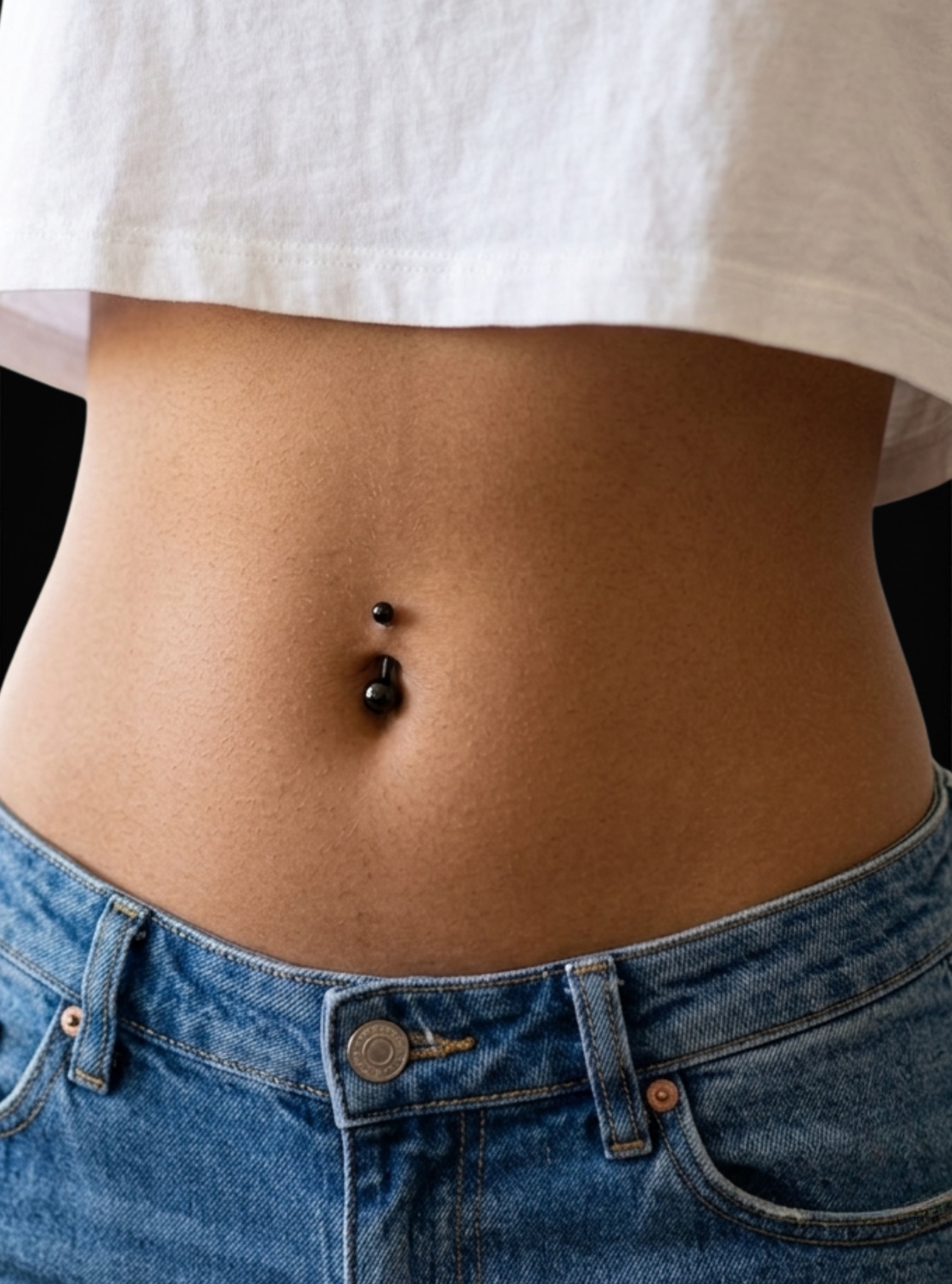 A close-up of a person's midsection showing a black double-stone belly button piercing, wearing a white t-shirt and blue jeans.