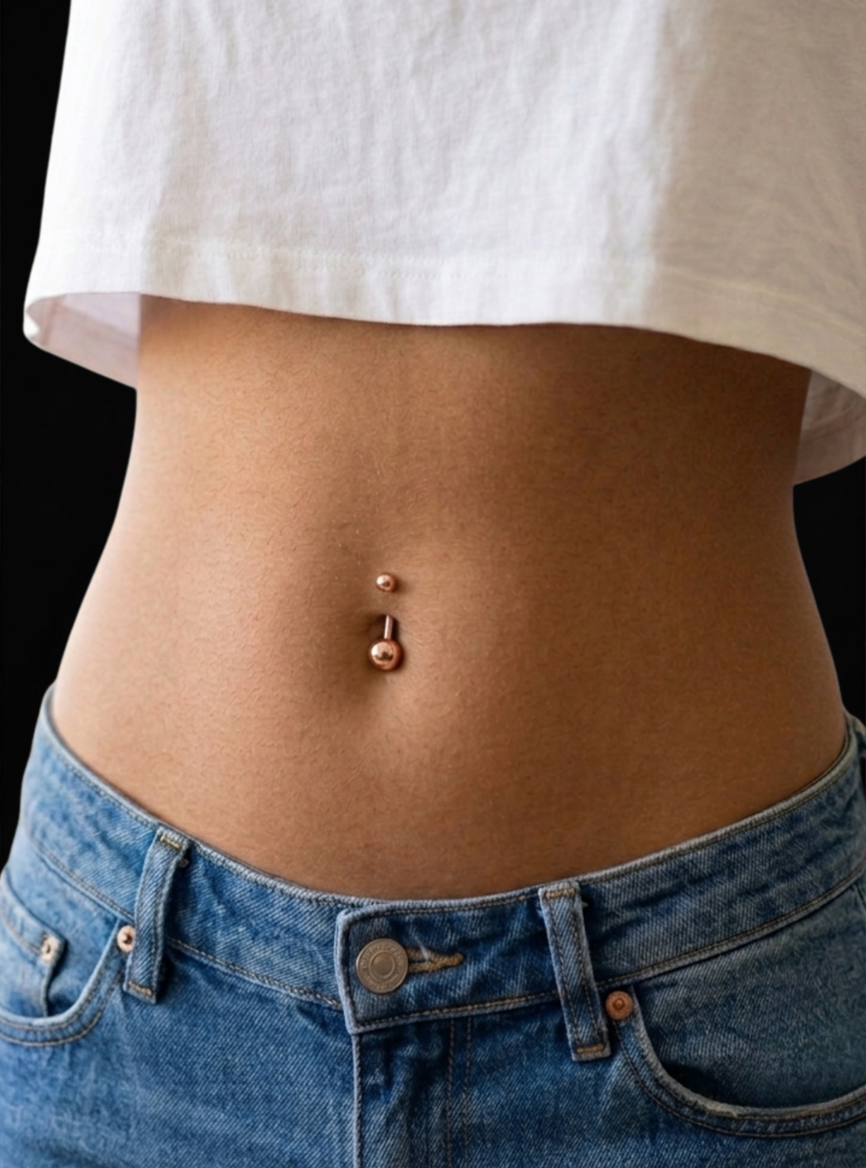 A close-up of a person's midsection wearing a rose gold navel piercing with a barbell and bead, paired with a white cropped t-shirt and blue jeans against a black background.