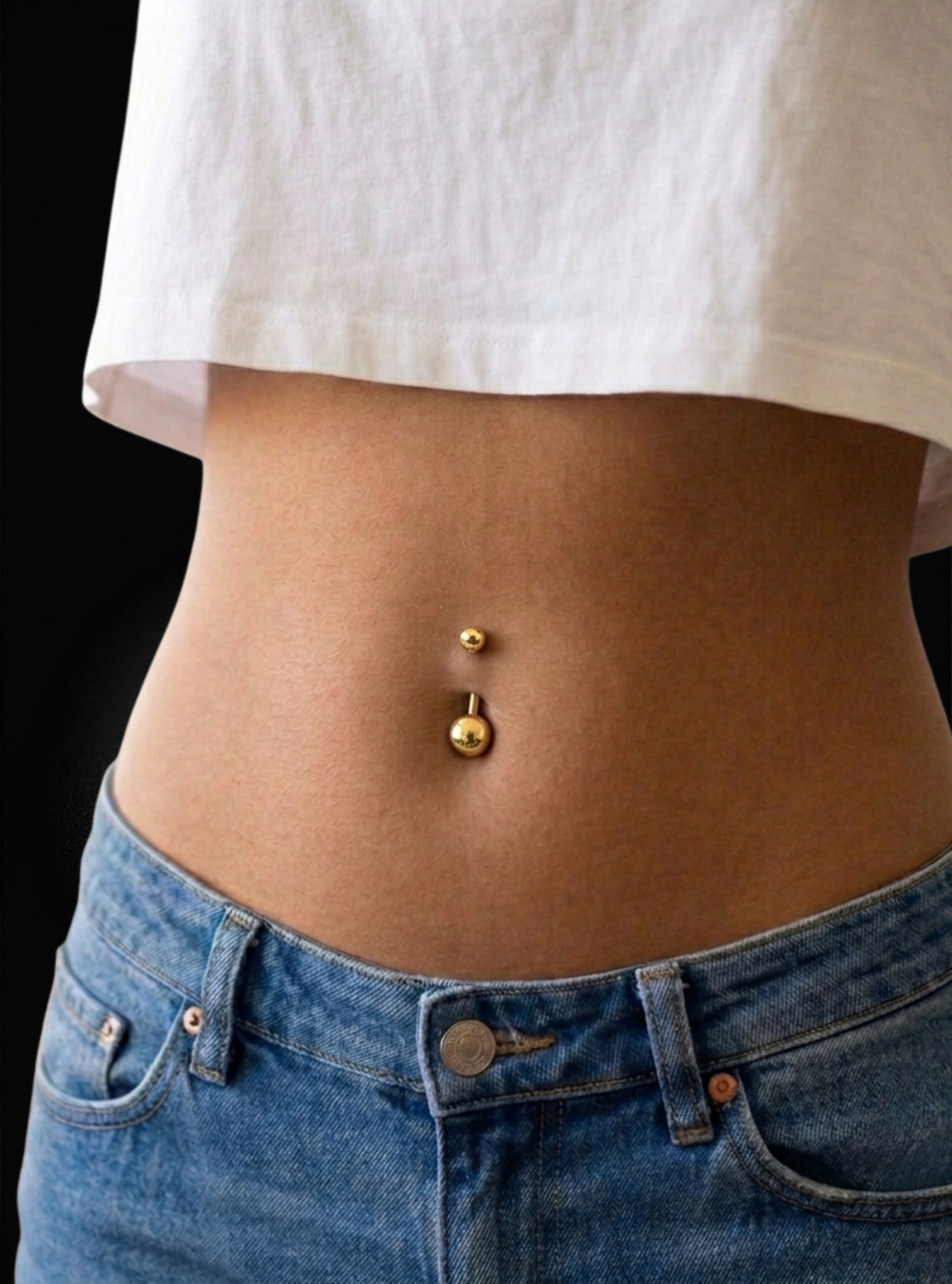 A close-up of a person's midsection wearing a gold belly button ring with a curved barbell and a small gold navel piercing, styled with a white crop top and blue jeans against a black background.