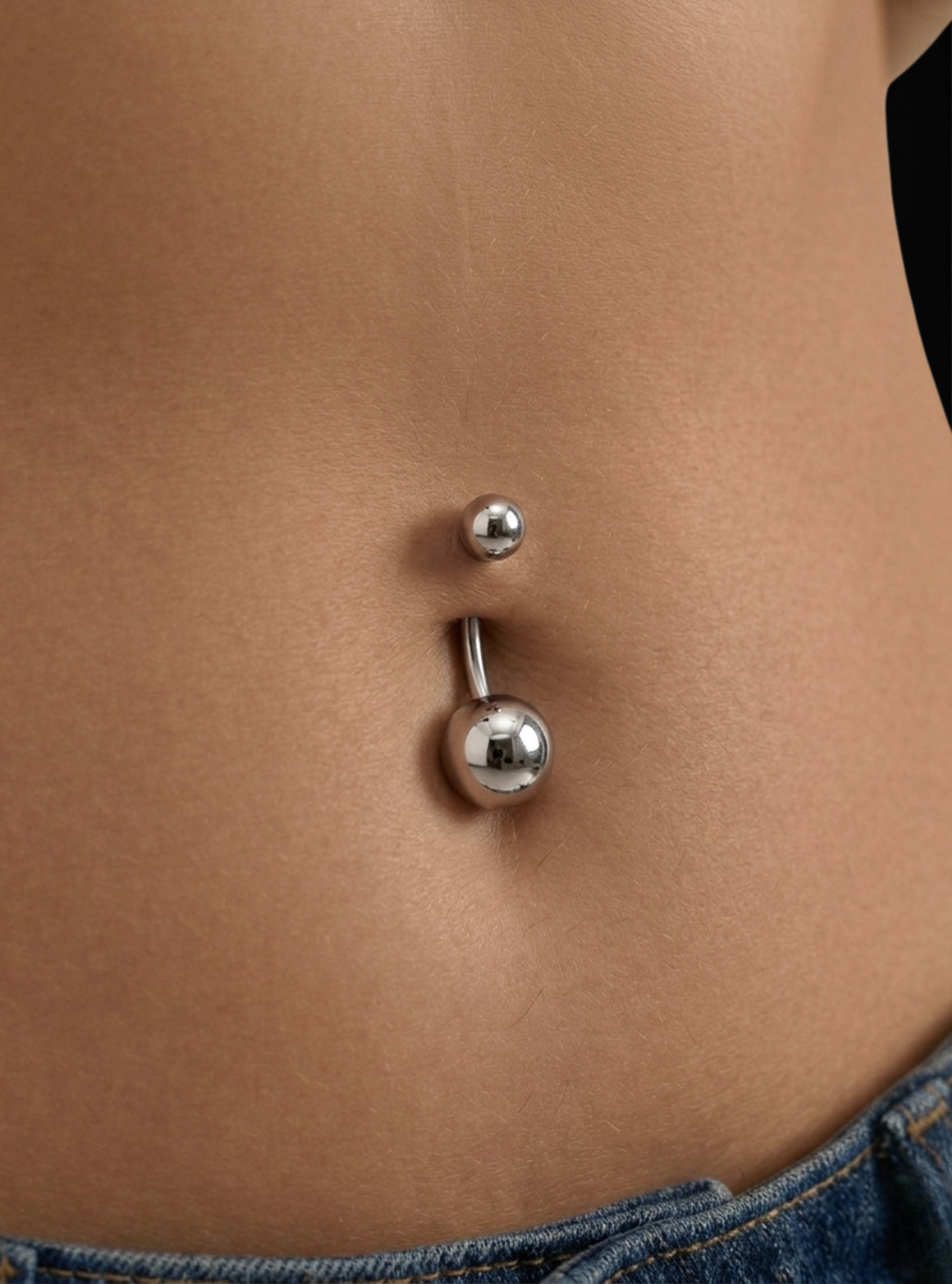 A close-up of a silver belly button ring with a small ball stud above and a larger ball stud below, worn on a person's navel with blue denim jeans visible at the bottom.