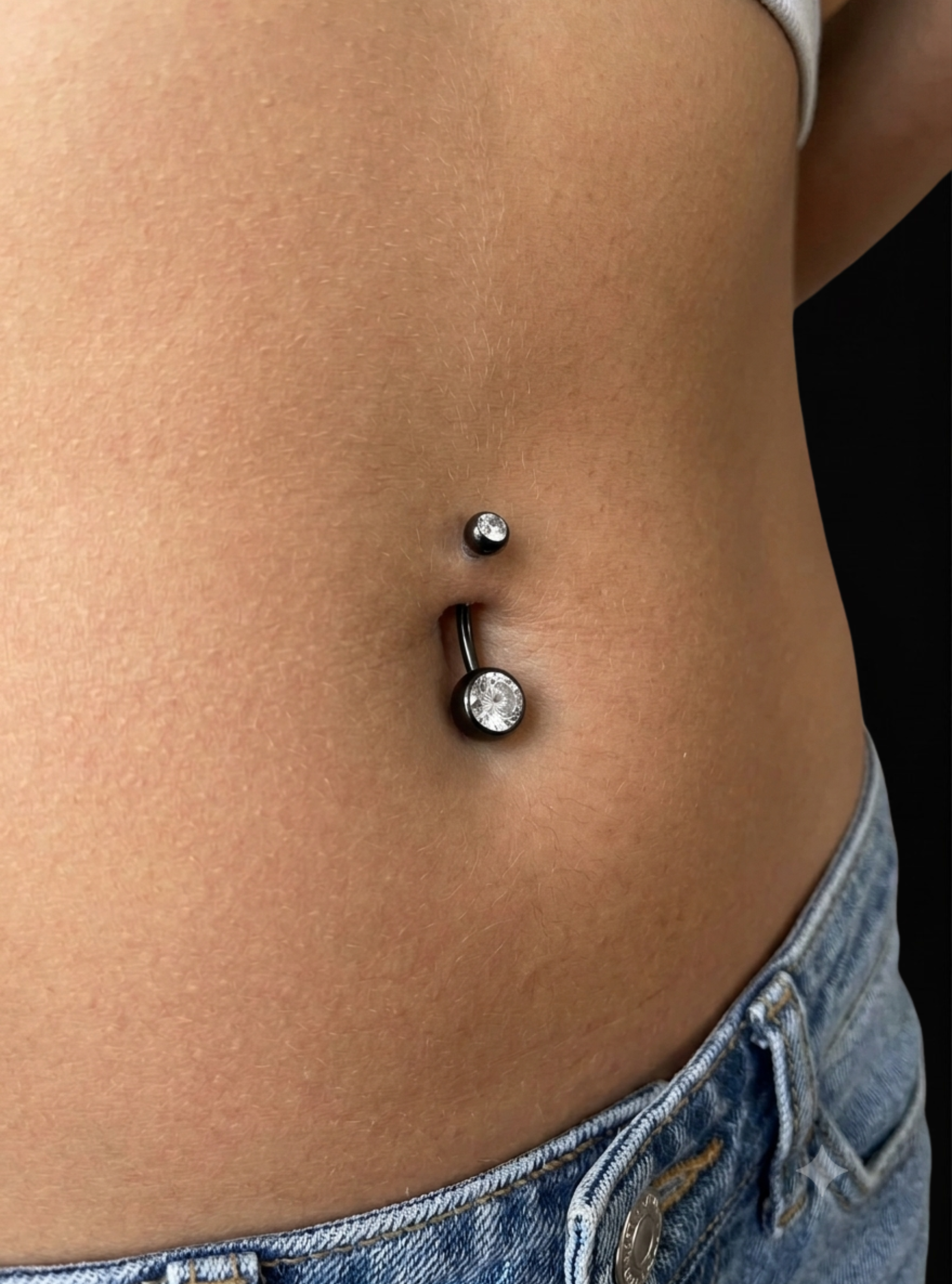 A close-up of a person's lower torso showing a belly button ring with a curved barbell design, one small silver stud above it, and a larger round clear rhinestone pendant below on a black bar.