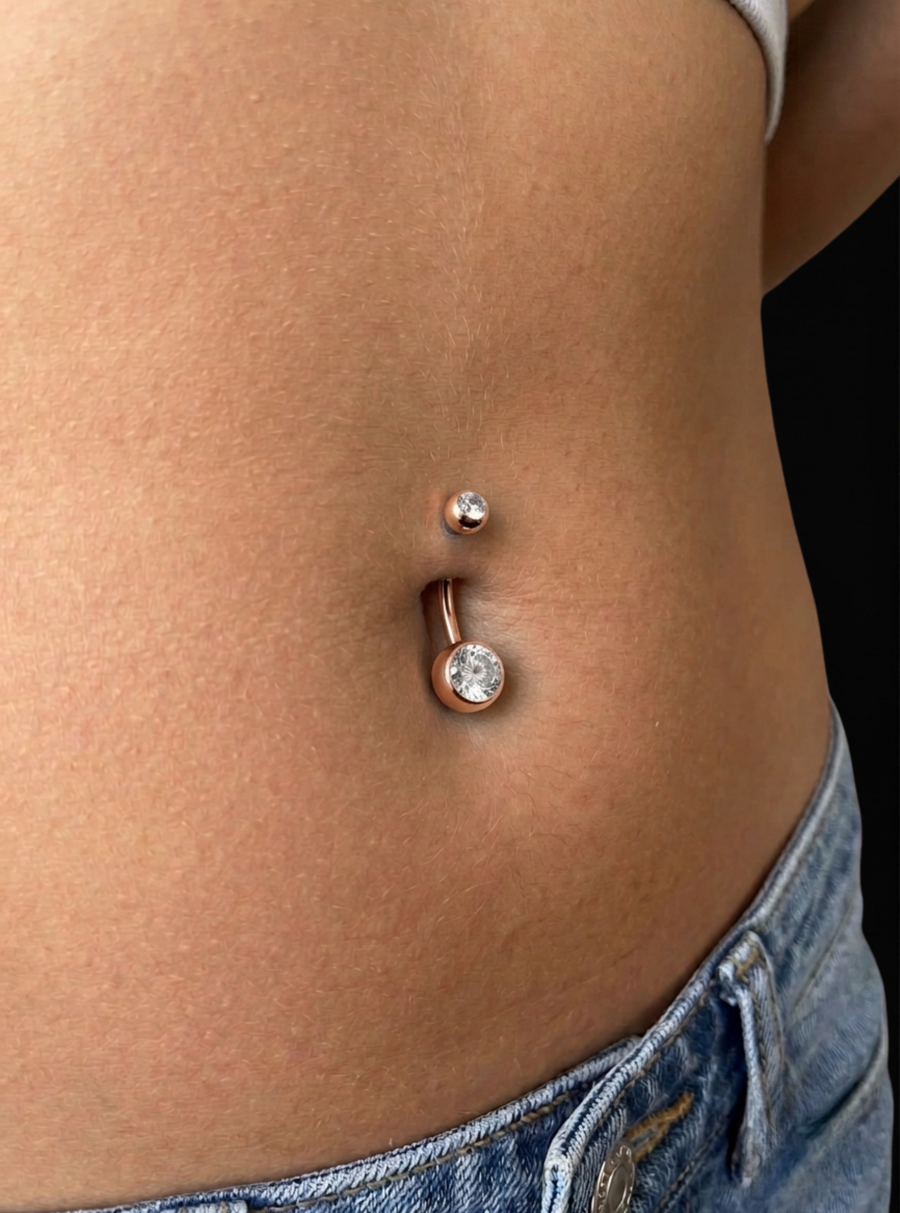 A close-up of a person's lower torso showing a rose gold curved barbell belly button ring with a clear rhinestone, worn with blue denim jeans against a black background.