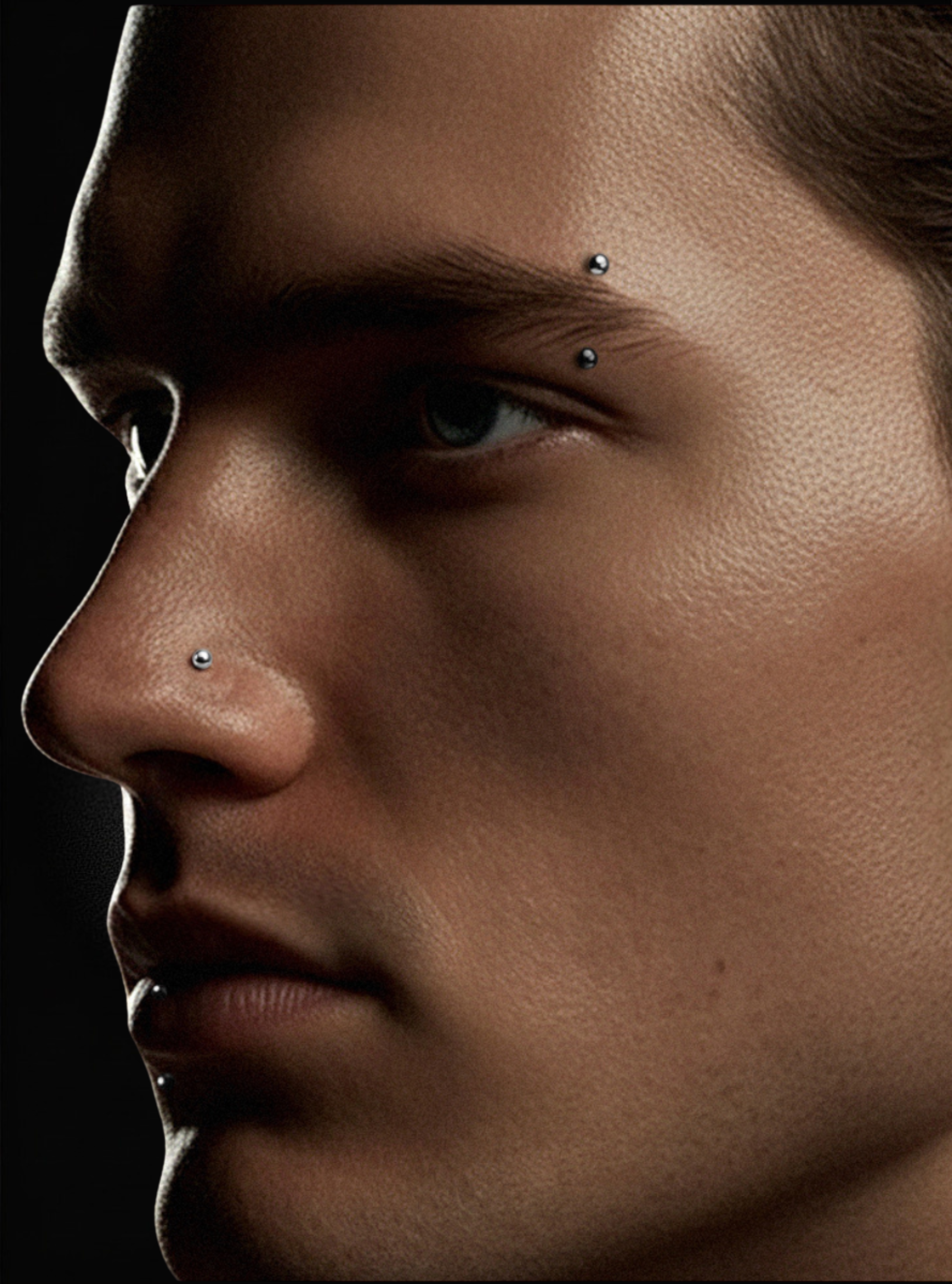 Close-up of a man's face in profile showing several small nose and eyebrow piercings against a dark background.itanium 