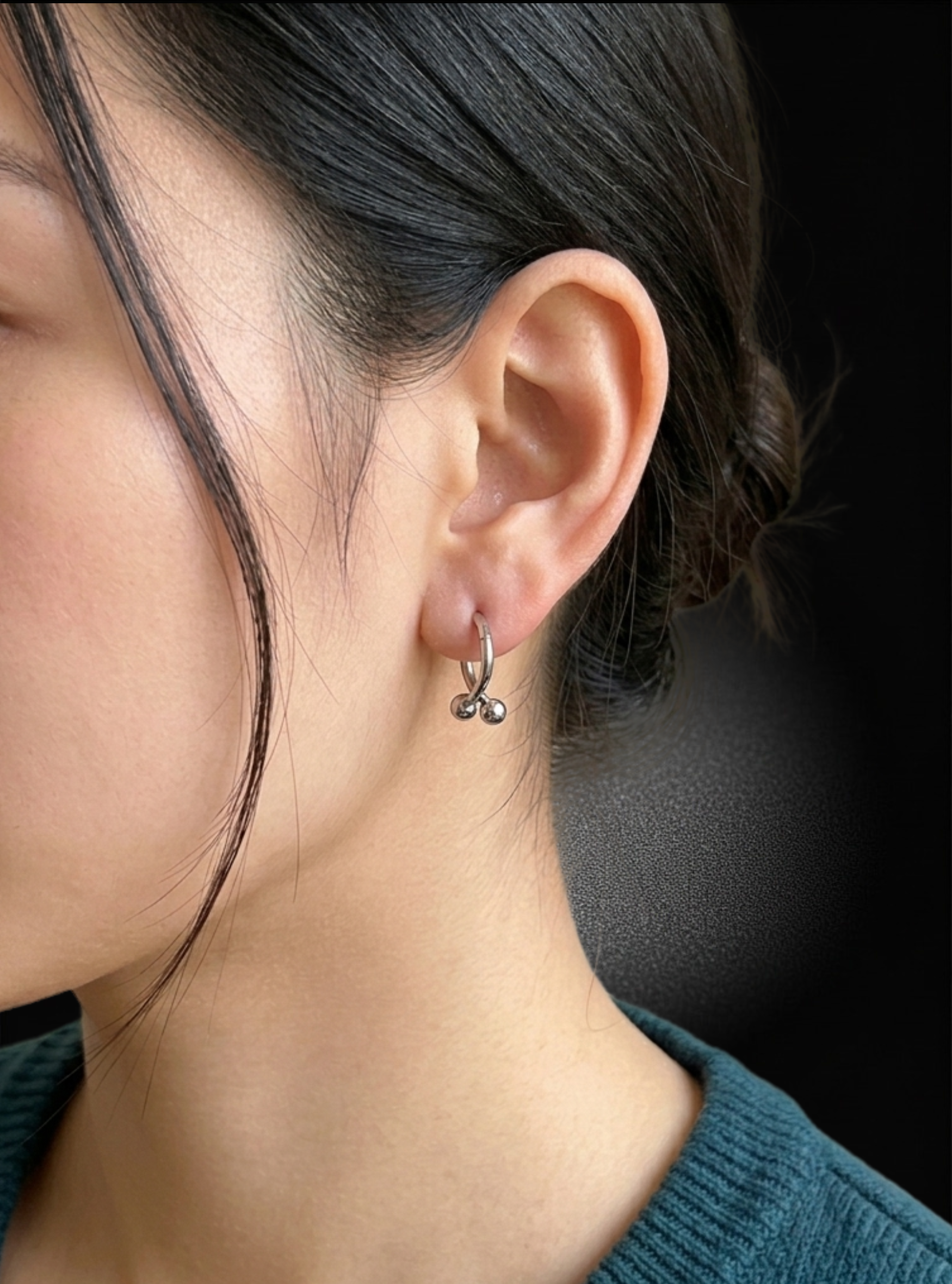 A close-up of a person's ear wearing a small silver hoop earring with two dangling bead-like charms, against a dark background.
