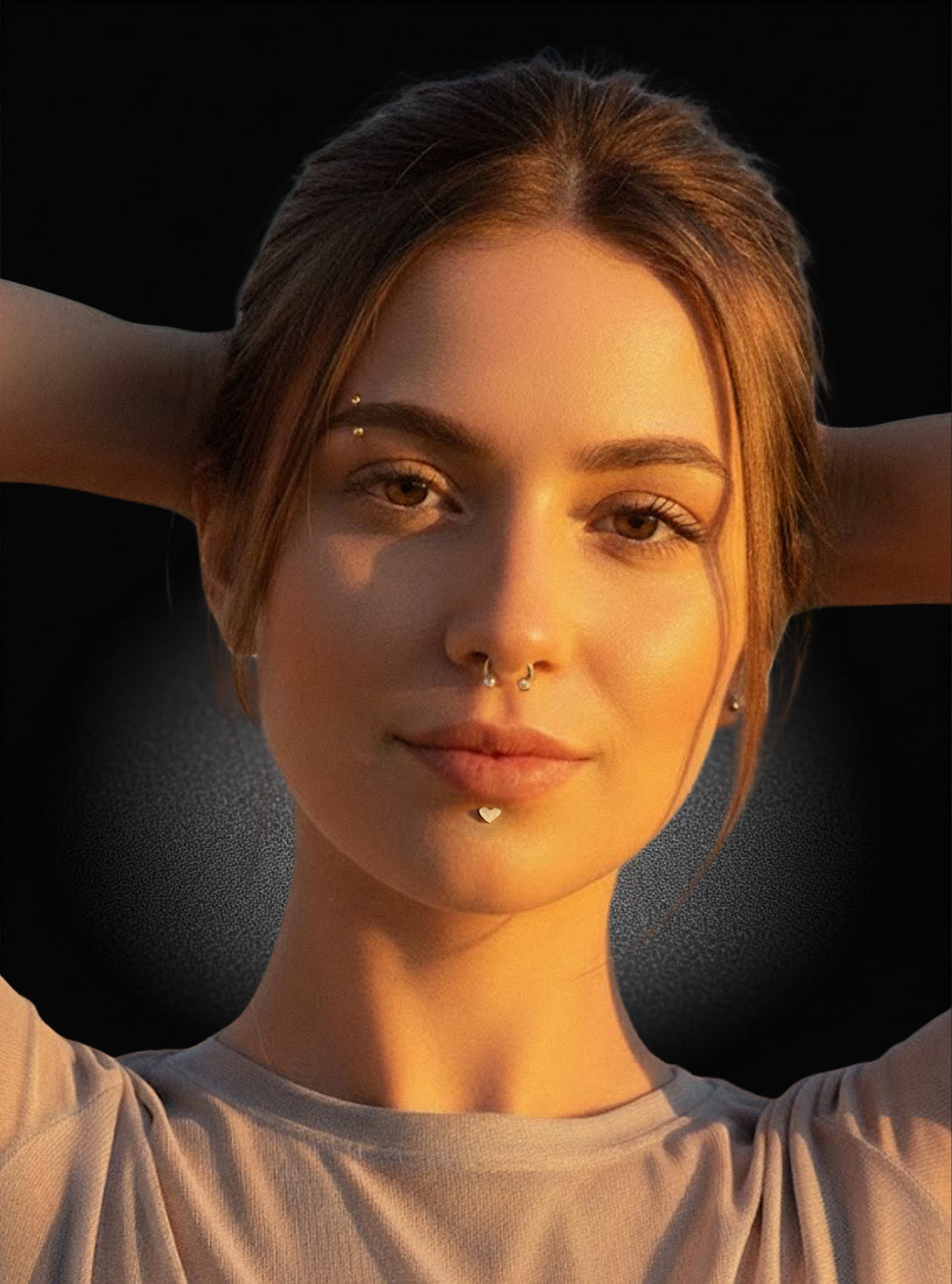 A close-up portrait of a woman with her hands behind her head, wearing a silver nose ring and a small lip piercing, set against a dark background with warm golden light.