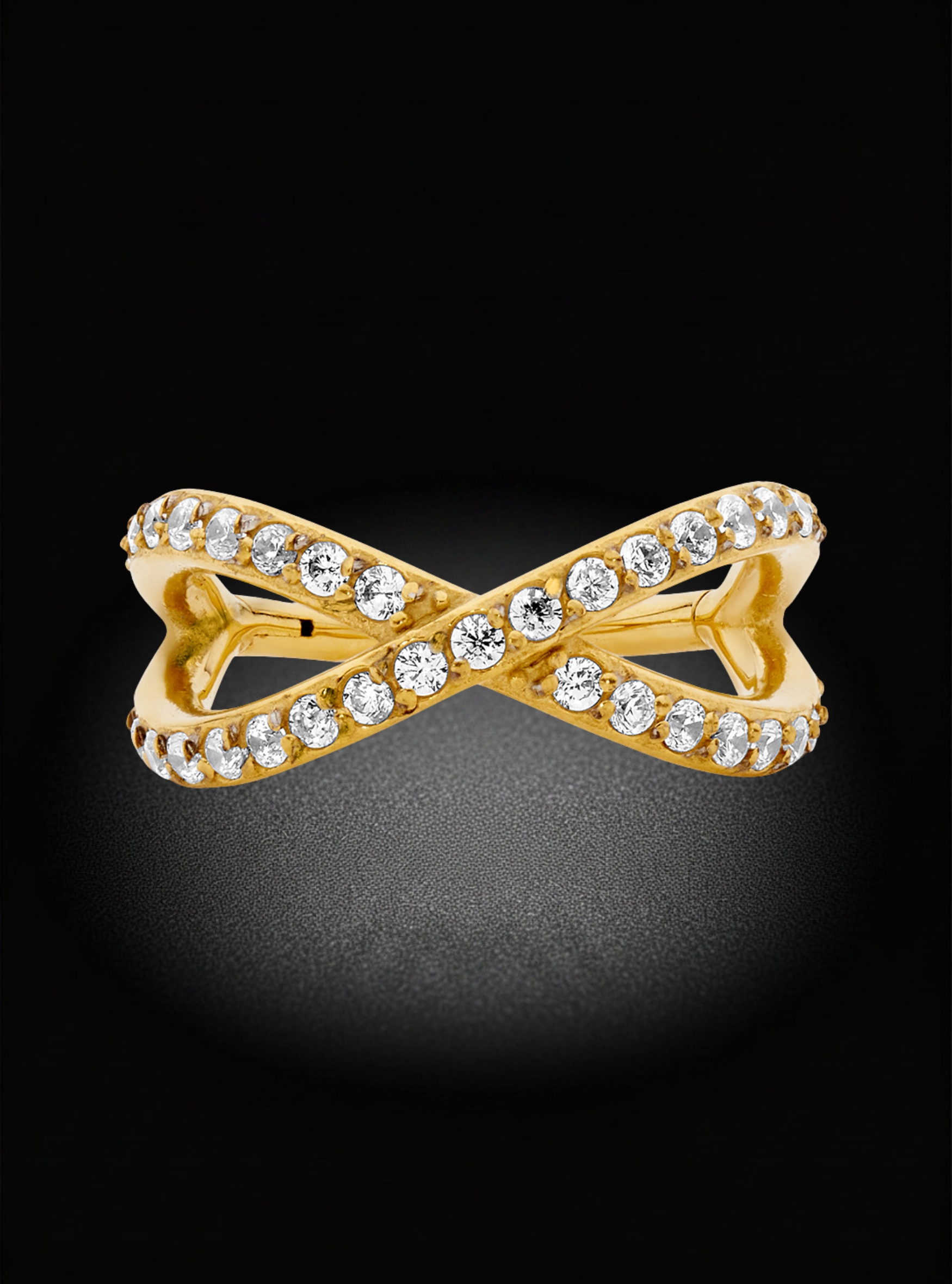 A yellow gold ring featuring an infinity-style double loop design, embellished with a row of clear round-cut stones along the top and bottom edges, set against a dark black background.