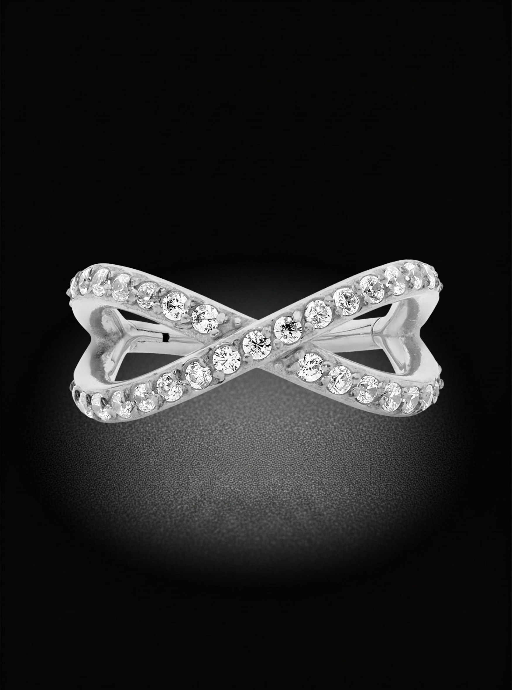 A silver-toned infinity-style ring set with clear round gemstones, photographed against a black background with a soft spotlight effect.