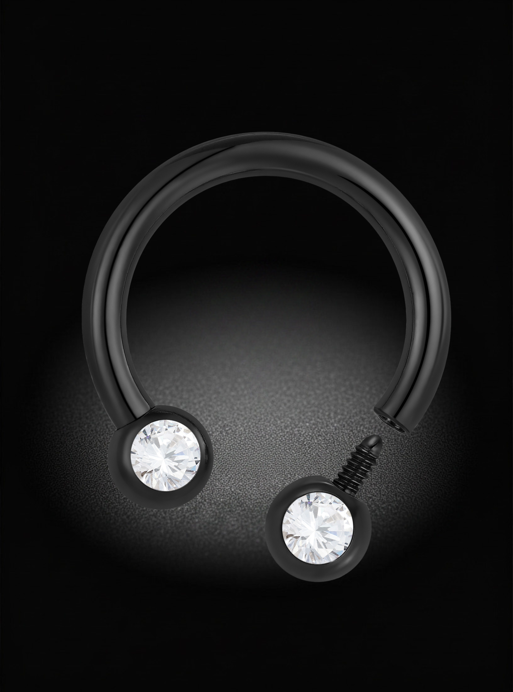 A black curved barbell-style body jewelry piece with two round bezel-set clear gemstones, photographed on a dark black background with a soft spotlight effect.