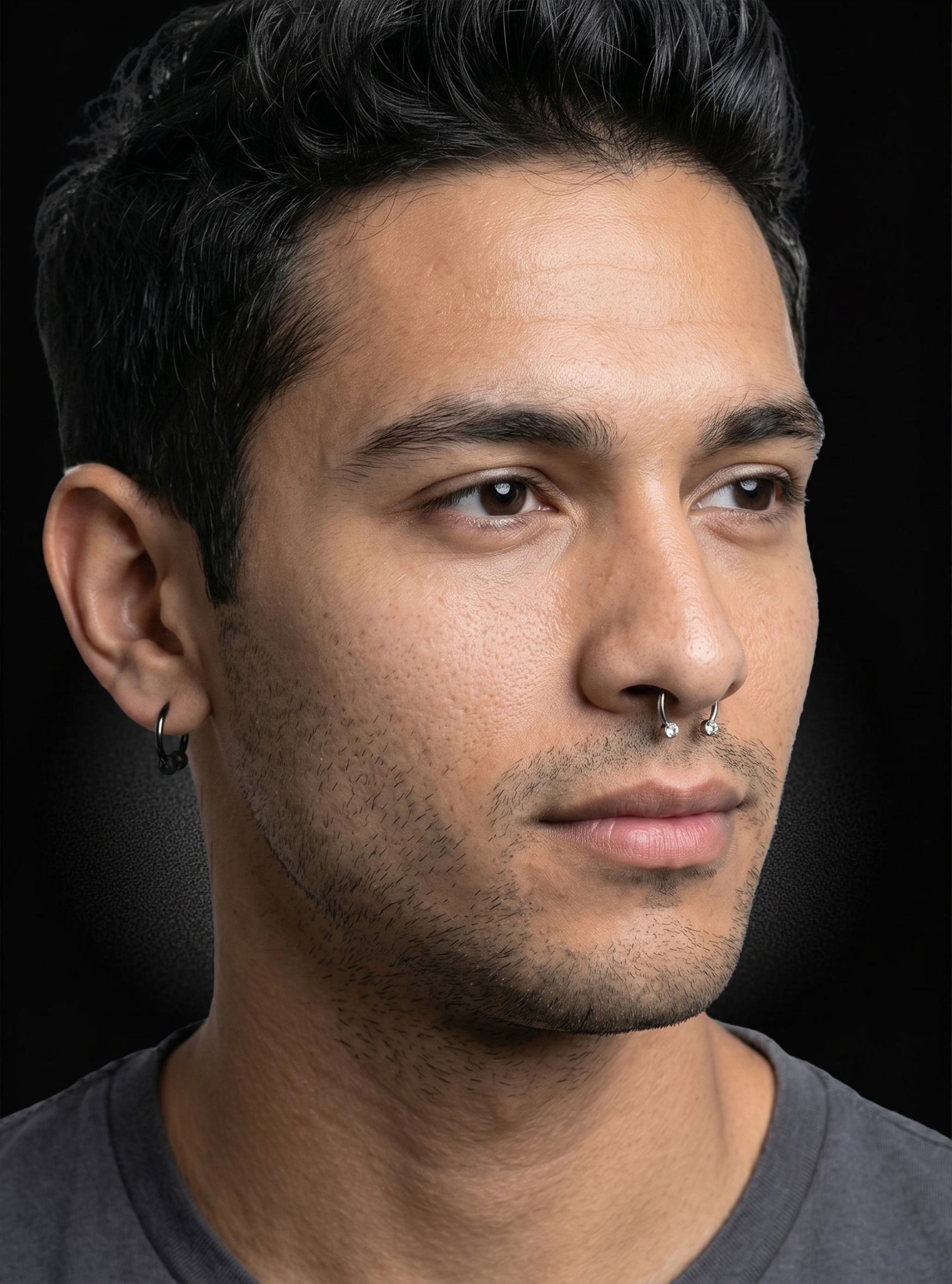 A close-up portrait of a man with dark hair, wearing a small black hoop earring in his left ear and a silver nose ring, against a dark background.