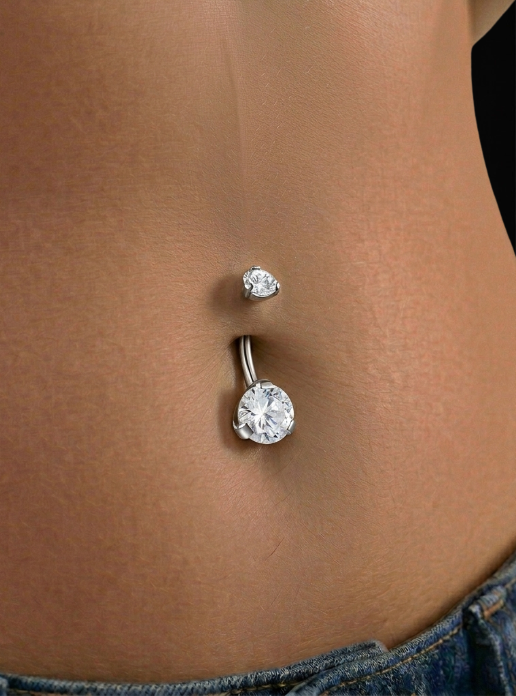 A close-up of a person's lower torso showing a silver-toned navel piercing with a large clear faceted gemstone and a smaller matching stud above it.