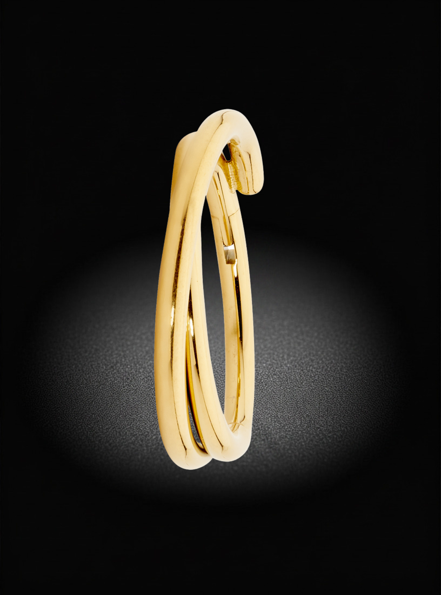 A polished yellow gold huggie hoop earring with a unique twisted, overlapping design, shown against a dark black background with a soft spotlight effect.