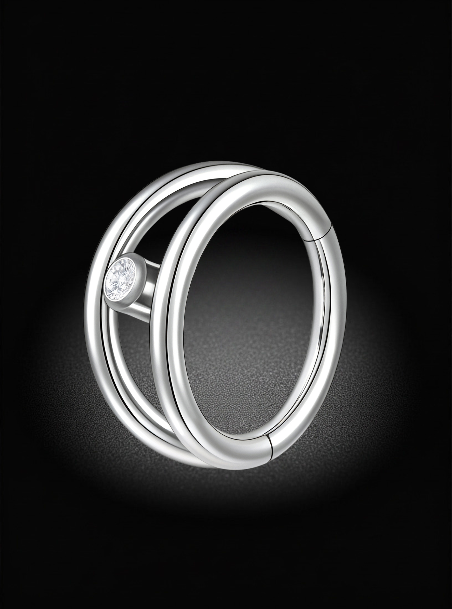 A polished silver-toned ring with a double-band design and a small clear gemstone accent, shown on a dark black background with a soft spotlight effect.
