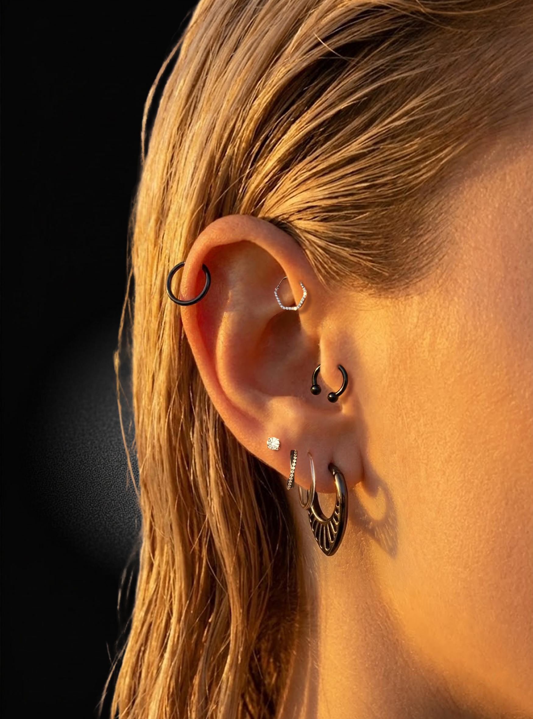 Close-up of a person's ear with multiple piercings wearing black and silver hoop, cartilage, and lobe earrings against a dark background.