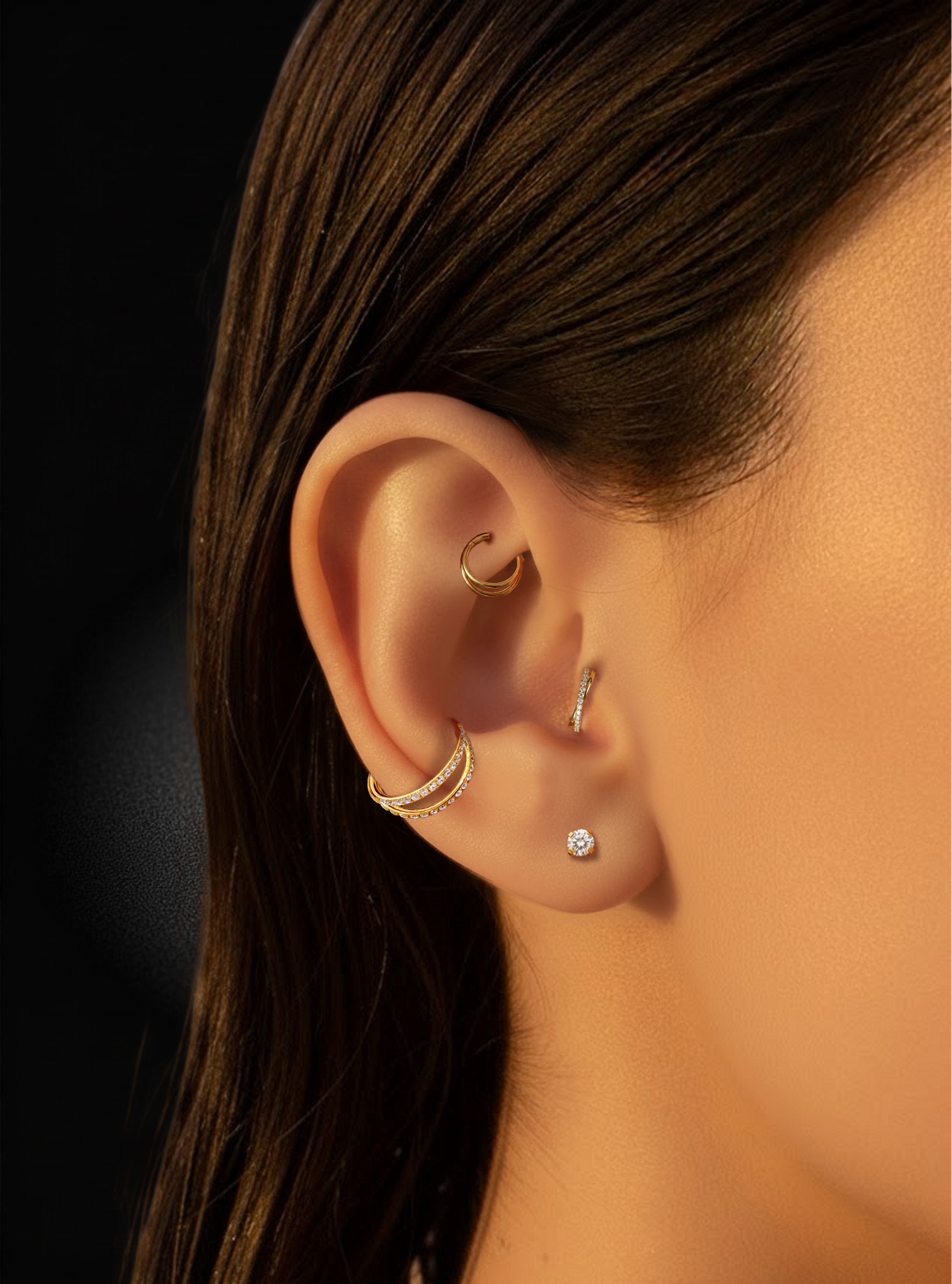 Close-up of a person's ear showcasing multiple gold and silver ear piercings, including a hoop in the cartilage, a curved barbell in the earlobe, and a small stud, set against dark hair and a black background.