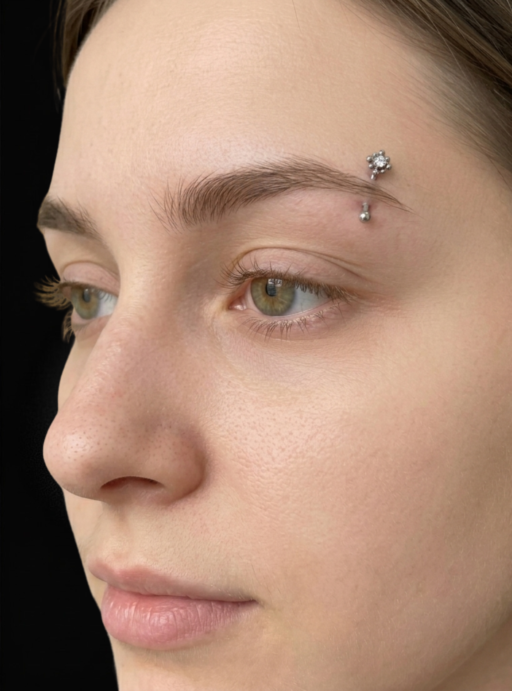 Close-up of a person's face showing eyebrow piercings with small titanium a barbell against a black background.flower cubic zirconia