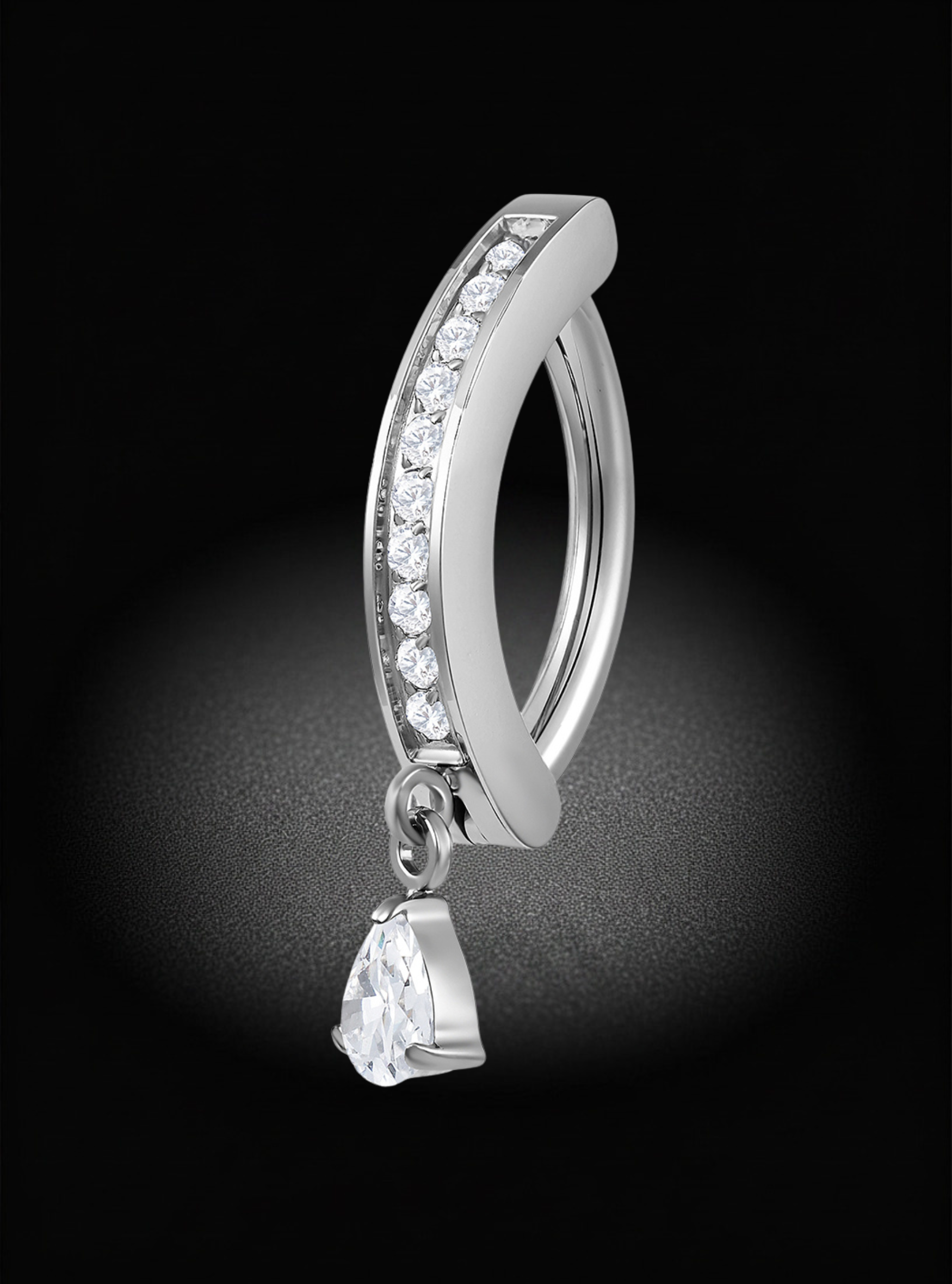 A silver-toned curved hinged clicker belly bar with a row of clear stones along the front and a dangling teardrop-shaped clear gemstone, shown on a black background with a spotlight effect.