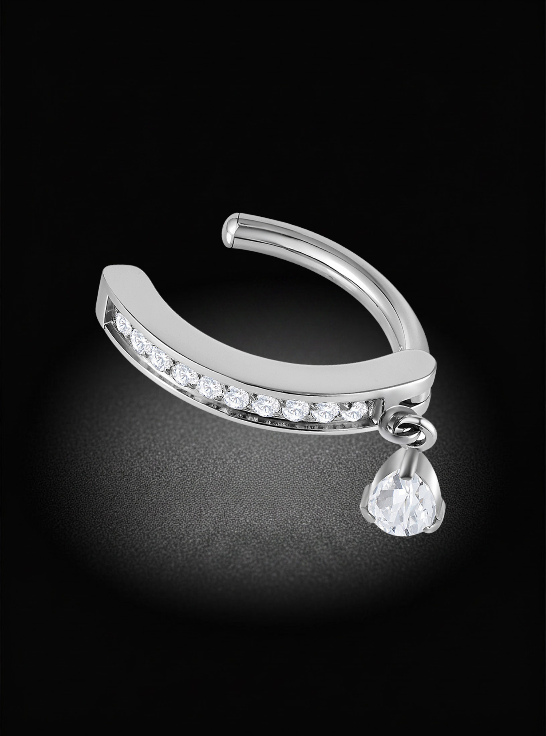 A silver-toned curved belly hinged clicker ring with a row of clear stones along the front and a dangling teardrop-shaped clear gemstone, photographed on a black background.