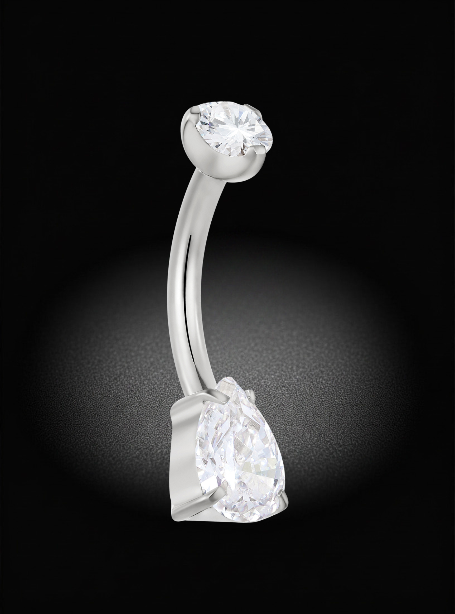 A curved silver-toned barbell belly button ring featuring a large clear teardrop-cut gemstone at the bottom and a smaller round-cut gemstone at the top, set against a dark background with a soft spotlight effect.