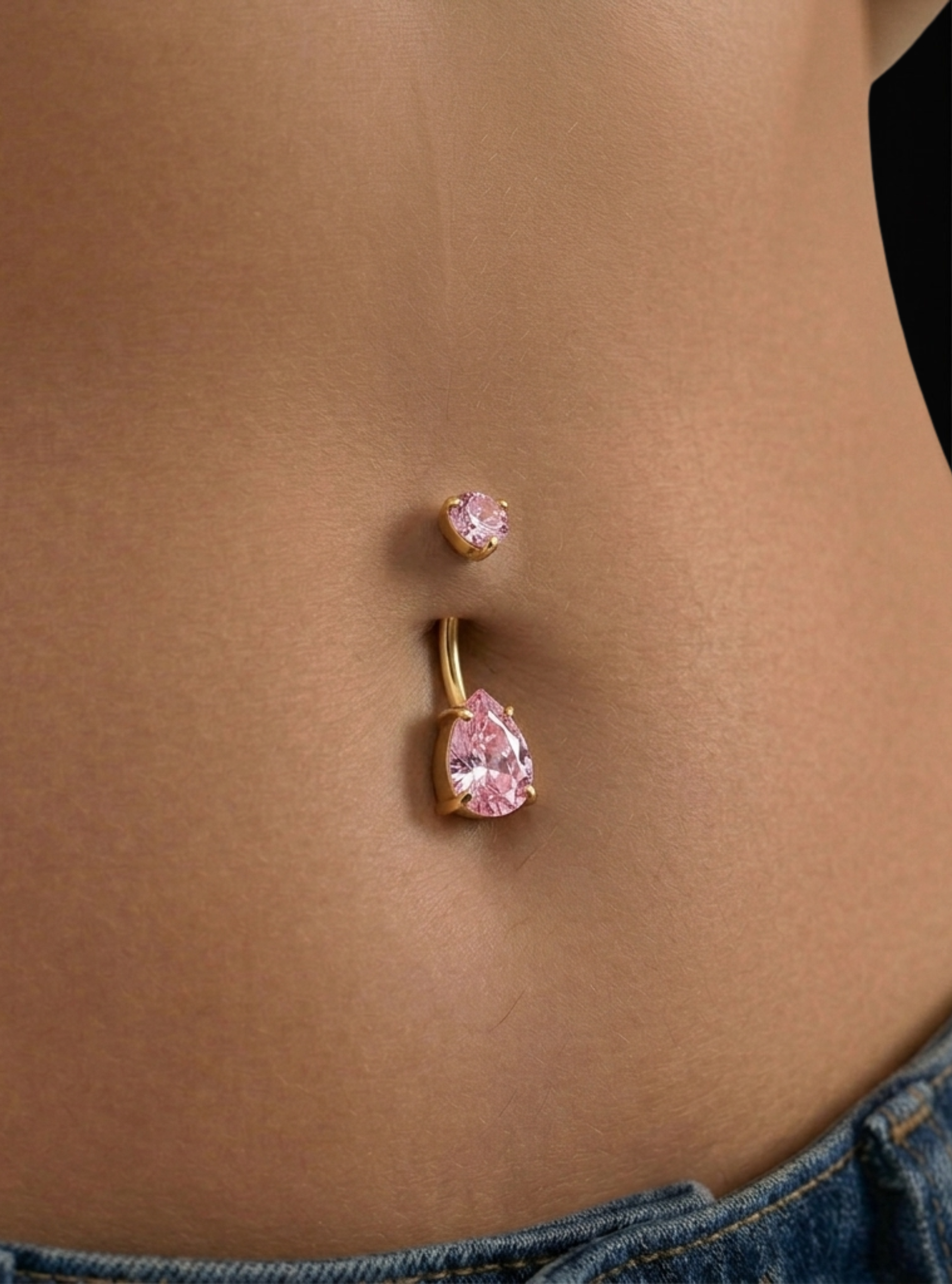 A close-up of a person's lower torso showing a gold belly button ring with a large pink teardrop gemstone and a smaller matching pink stud above it.