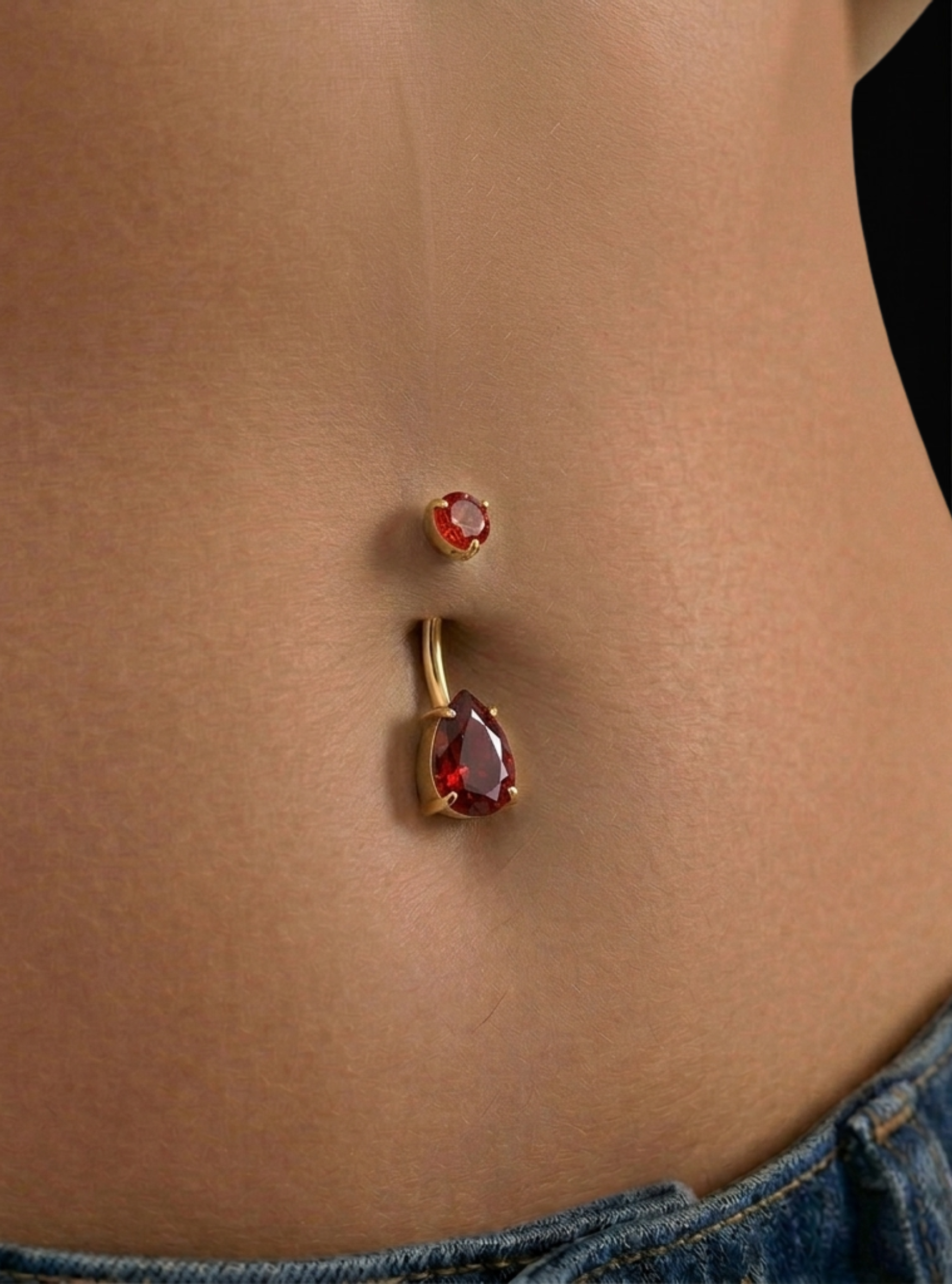 Gold belly button ring with a teardrop-shaped red gemstone and a matching small red gemstone stud, shown worn on a person's abdomen.