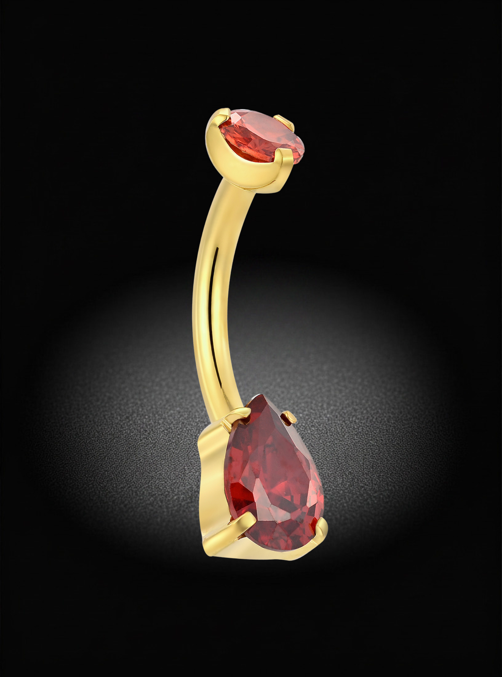 A curved gold barbell belly button ring featuring a large teardrop-shaped red gemstone on one end and a smaller oval red gemstone on the other, set against a dark background with a spotlight effect.