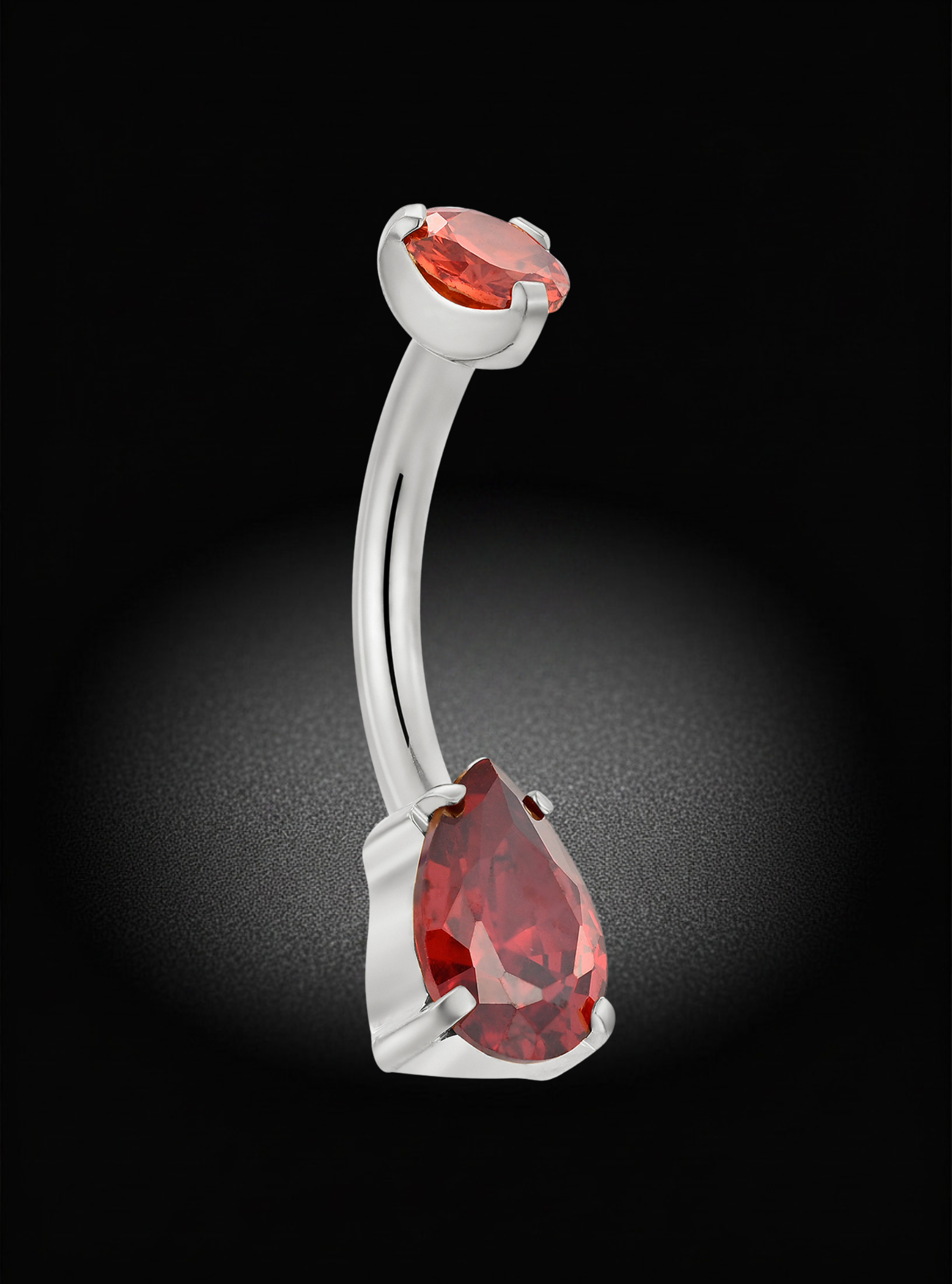 A curved barbell belly button ring featuring a large faceted red teardrop gemstone and a smaller oval red gemstone set in a polished silver-tone metal, shown against a dark background with a spotlight effect.
