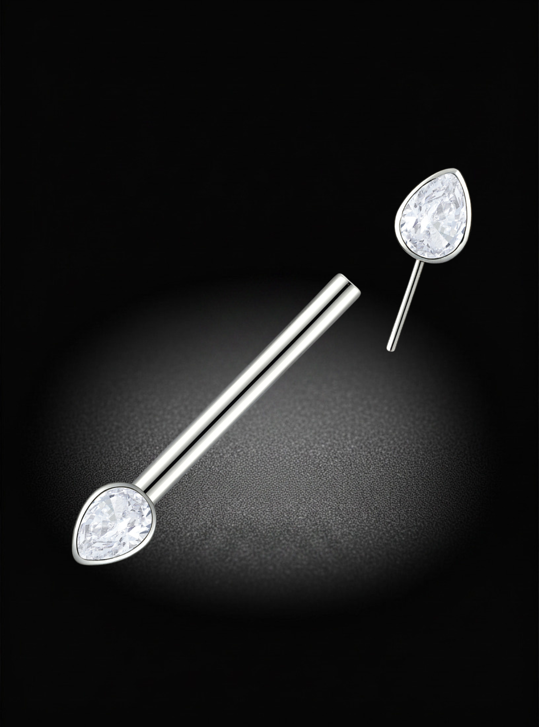 A pair of silver titanium nipple barbells with teardrop-shaped clear gemstones, shown on a black background with a spotlight effect.