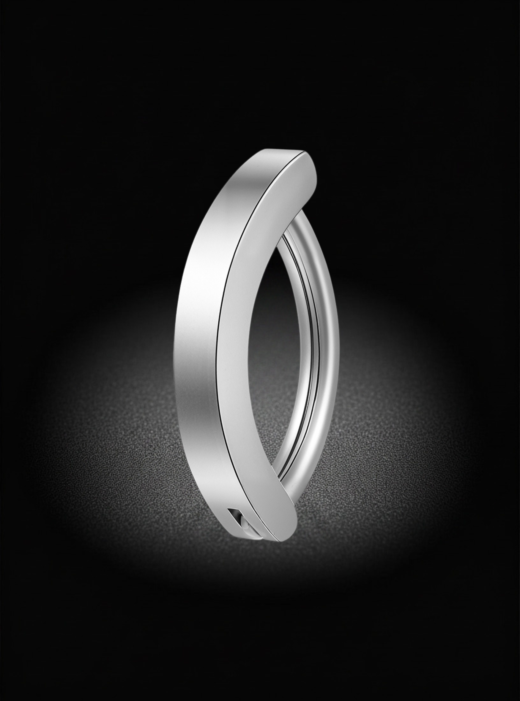 A minimalist, polished silver-toned metal ring with a unique curved, open-ended design, shown on a dark background with a soft spotlight effect.