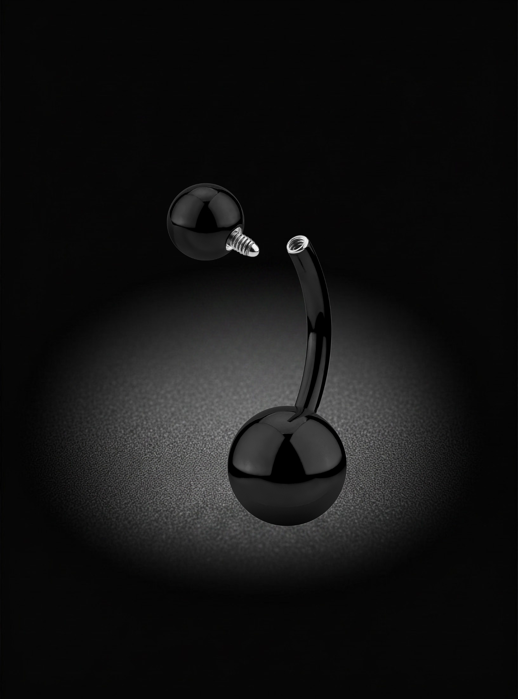 A black curved belly barbell piercing jewelry piece with two spherical ball ends and a screw-back top, shown against a dark background with a spotlight effect.