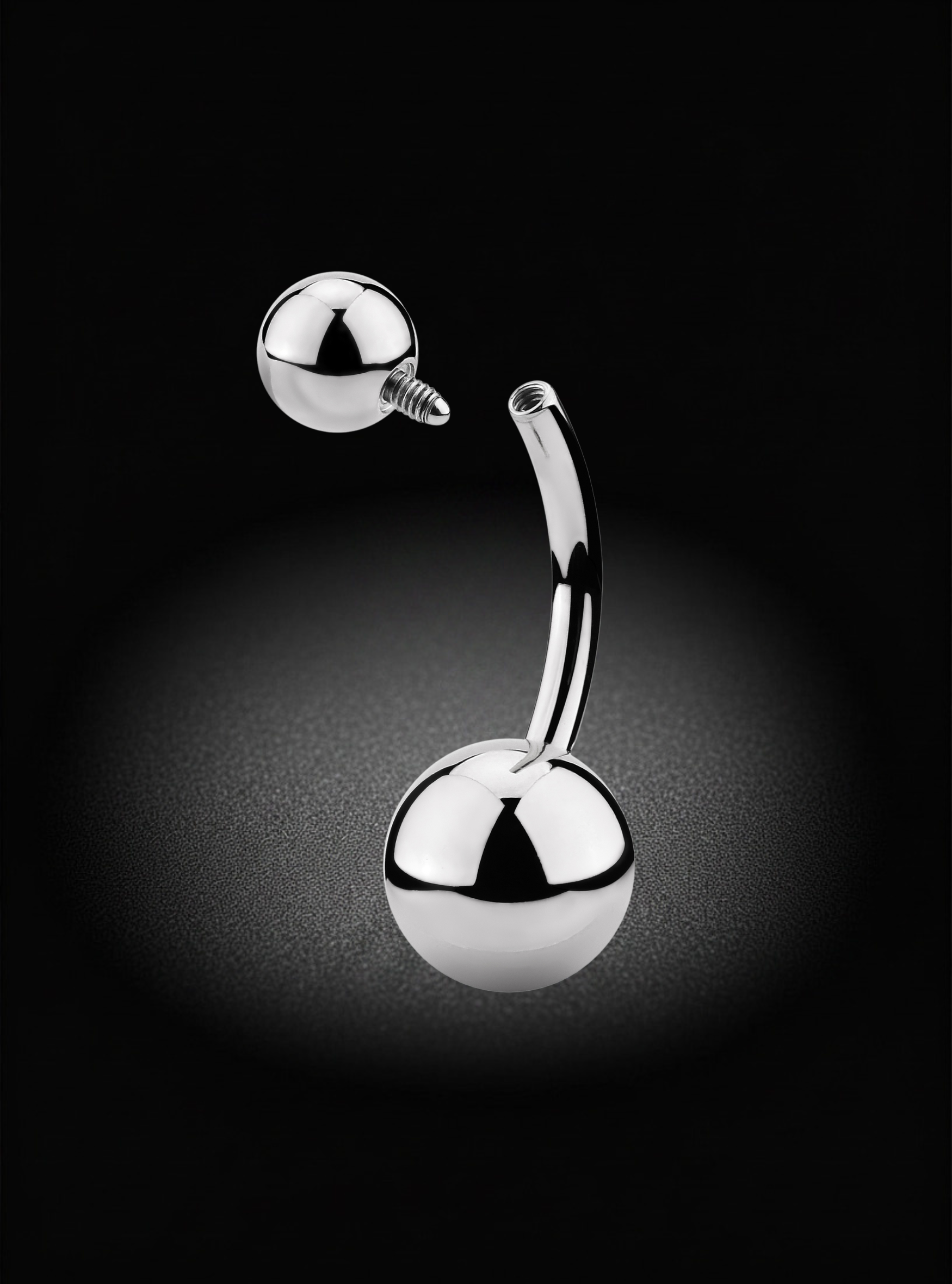 A curved stainless steel barbell belly button piercing with two polished ball ends, shown against a dark black background with a soft spotlight effect.