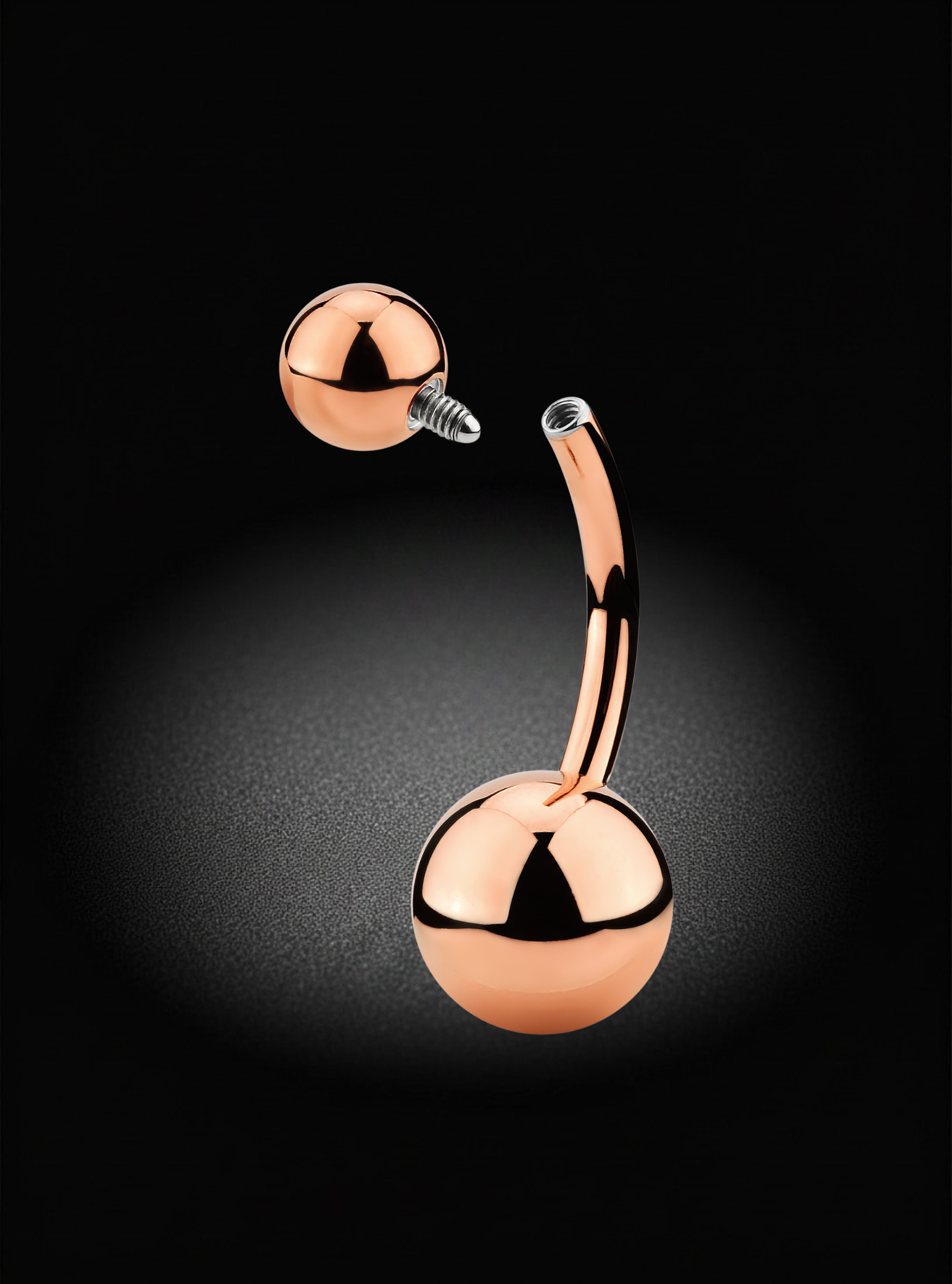 A curved barbell belly button piercing with two polished rose gold ball ends, one of which is shown detached to display the screw-back post, set against a dark spotlighted background.
