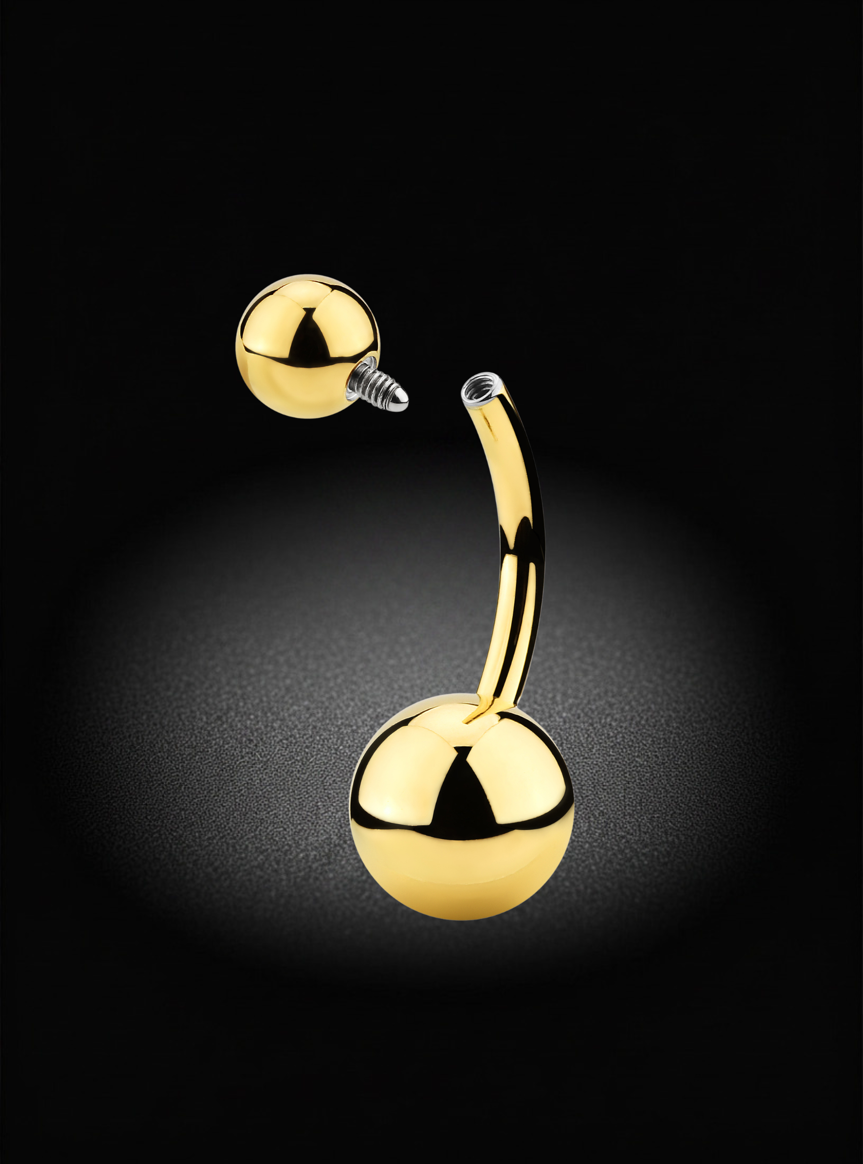 A curved barbell belly button piercing stud with polished gold-tone spheres on both ends, shown against a dark black background with a soft spotlight effect.