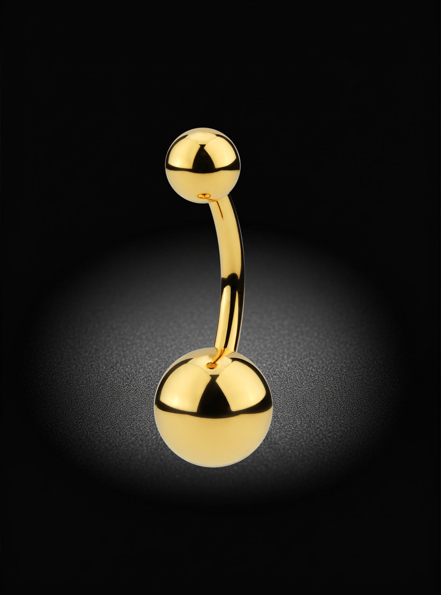 A curved barbell belly button piercing with polished gold-tone spheres on both ends, shown against a black background with a soft spotlight effect.