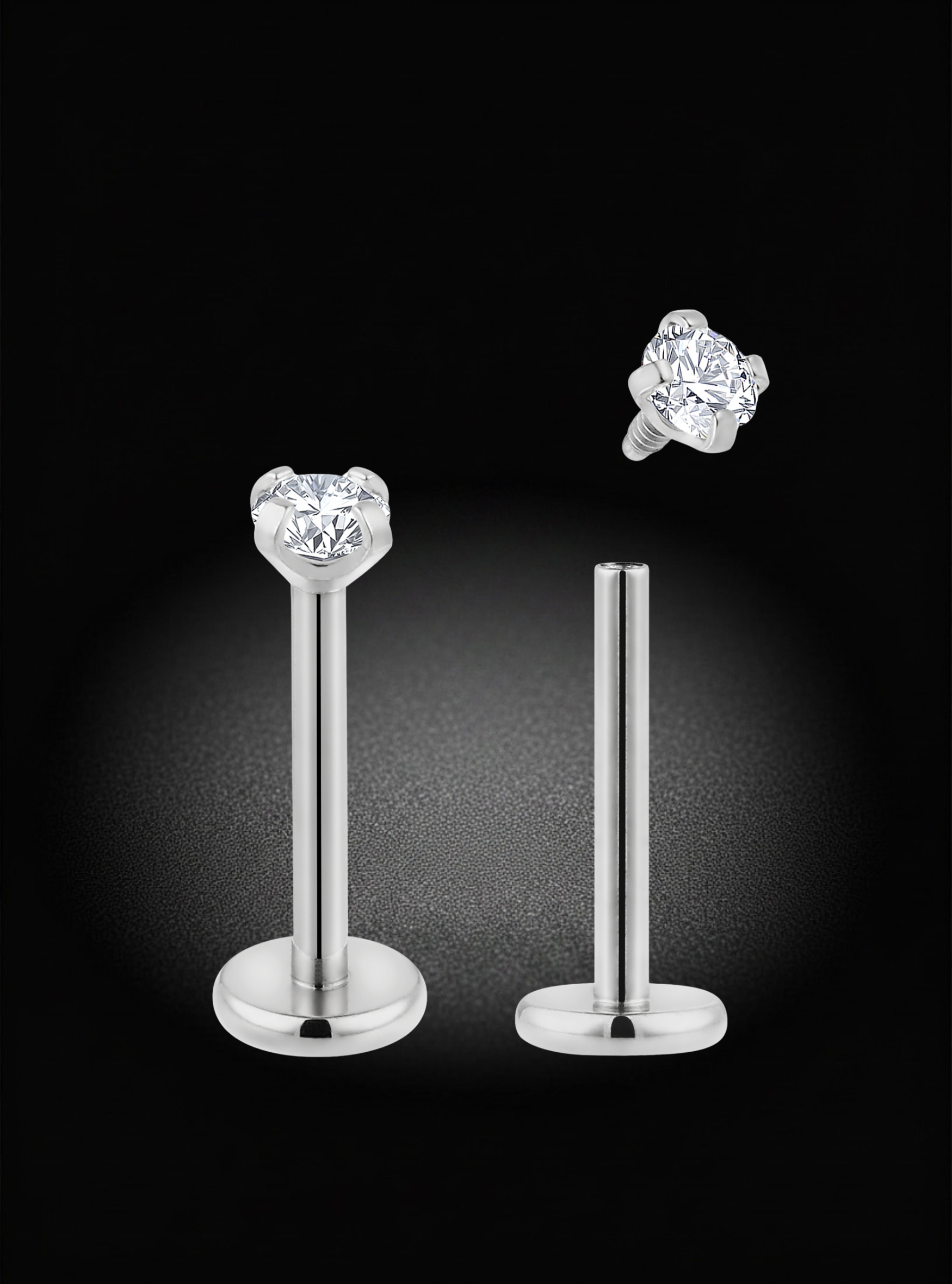 A pair of silver-toned labret earrings with heart-shaped clear gemstones, shown on a black background with one earring displayed front-facing and the other floating to show the back screw-back setting.