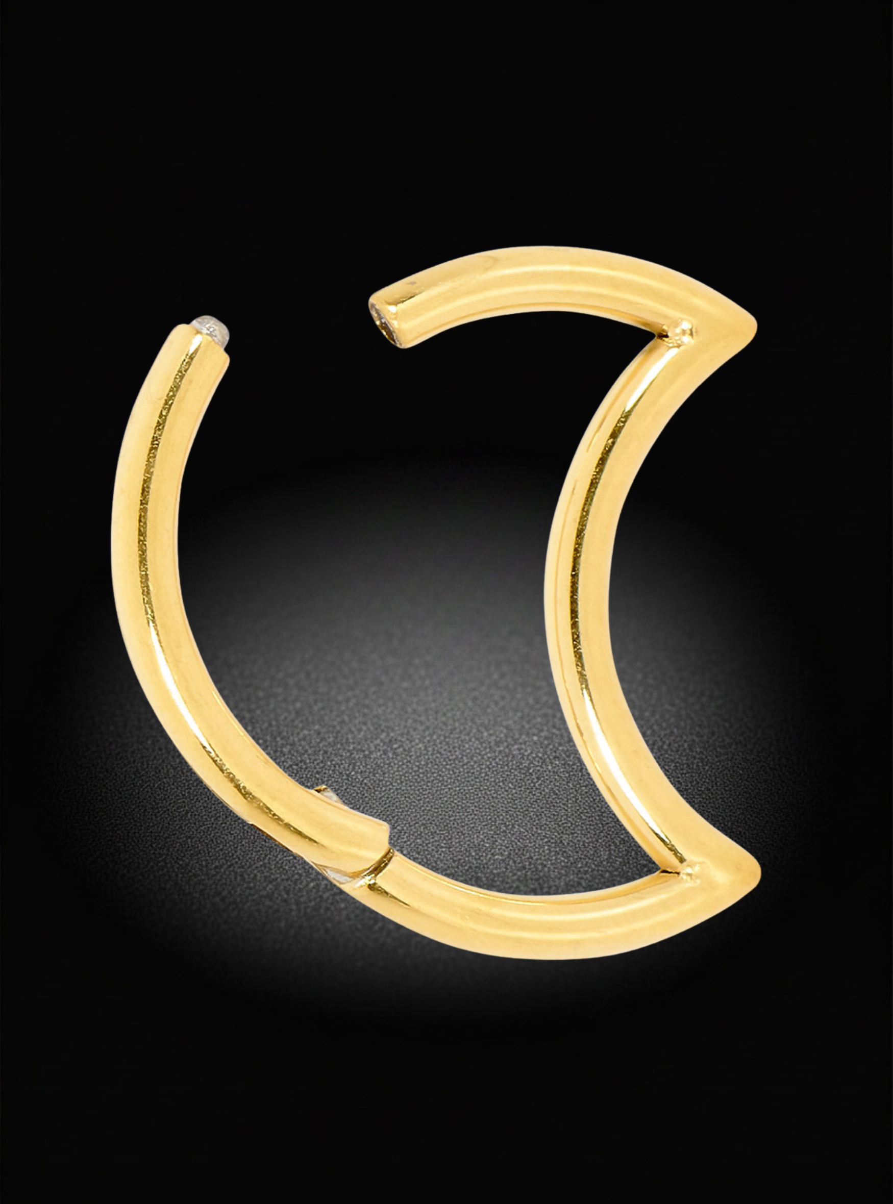 A polished gold crescent-shaped hoop earring with a small clear gemstone on the left tip, displayed on a black background with a soft spotlight effect.