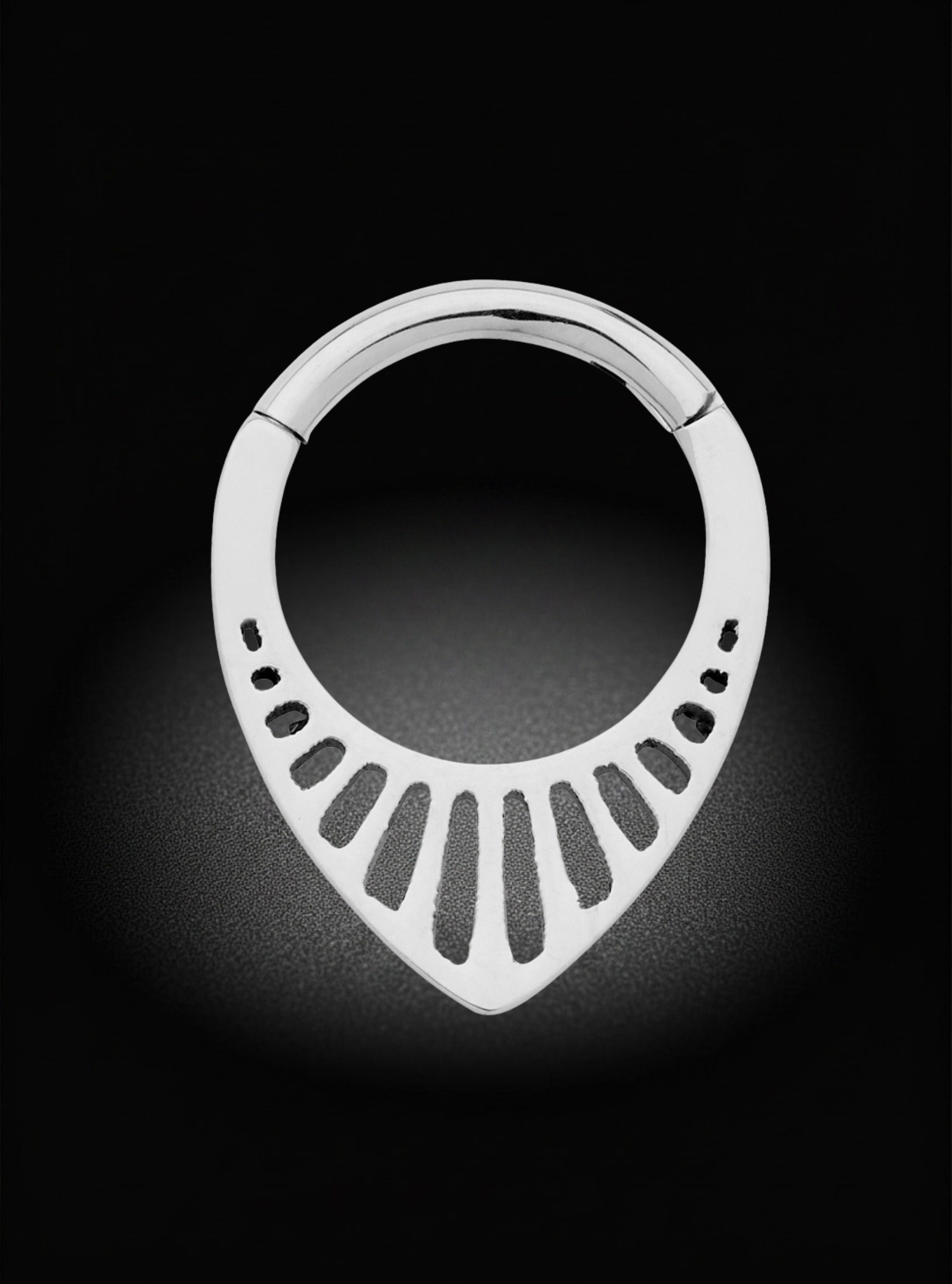 A silver metal ring with a teardrop-shaped cutout design and small oval details along the lower curve, shown against a black background with a soft spotlight effect.