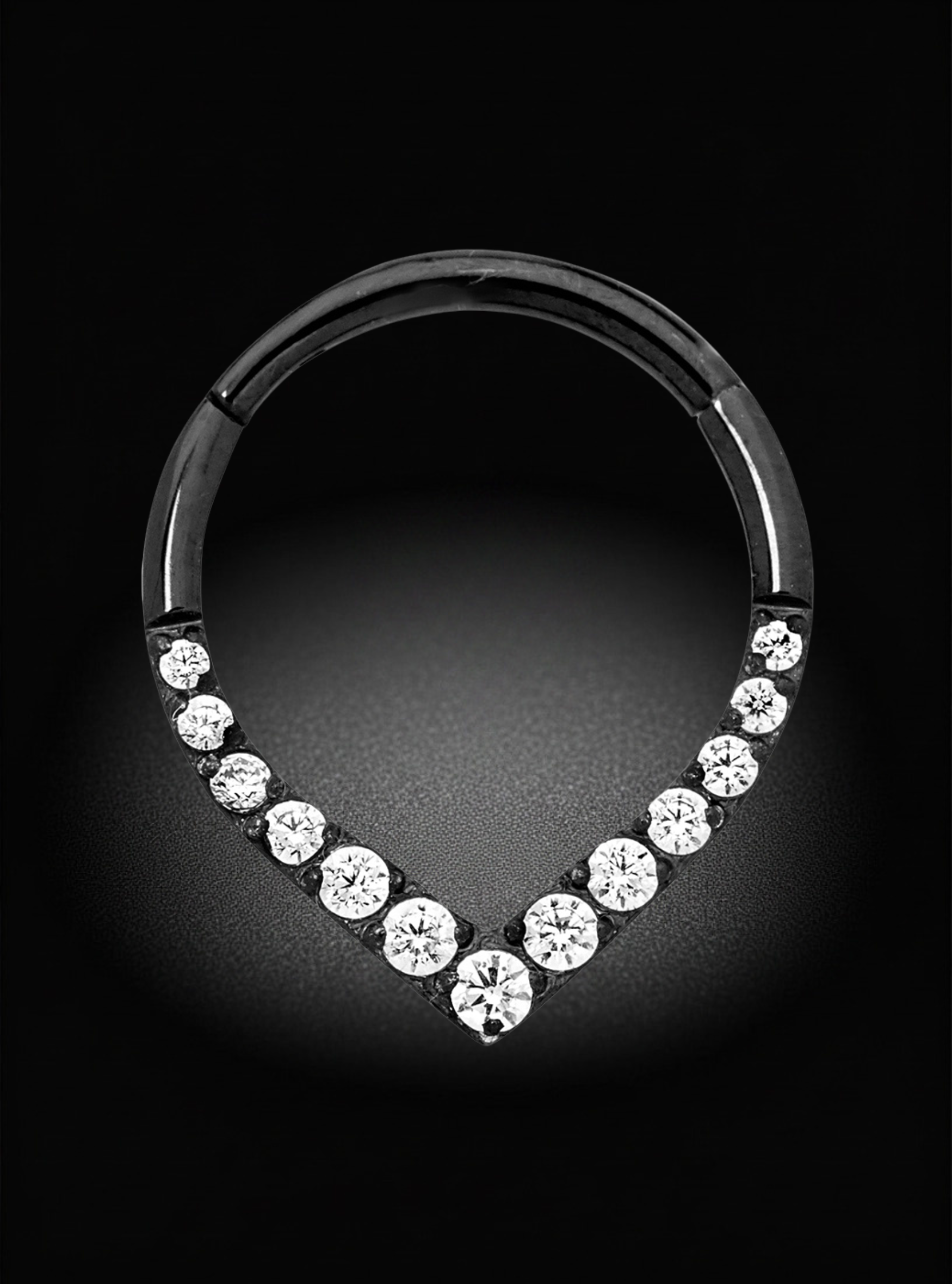 A silver-toned hoop piercing ring with a V-shaped row of clear round gemstones at the bottom, photographed on a black background.