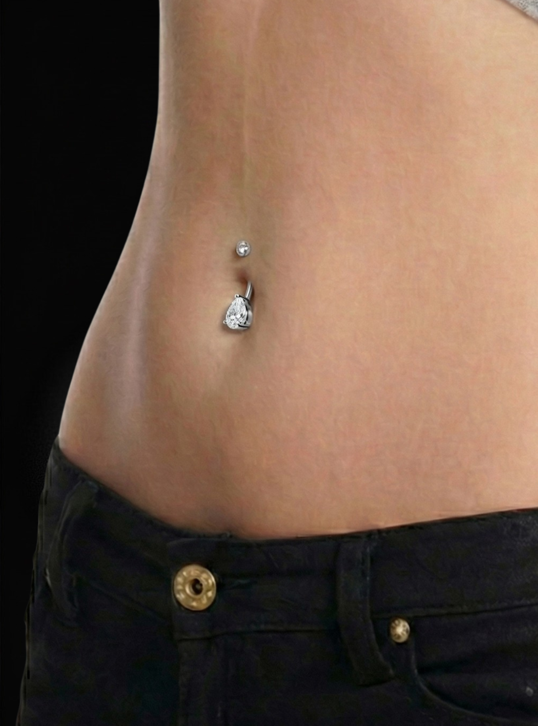 A close-up of a person's lower torso showing a silver belly button ring with a teardrop-shaped clear rhinestone and a small silver bead, worn with black jeans against a black background.