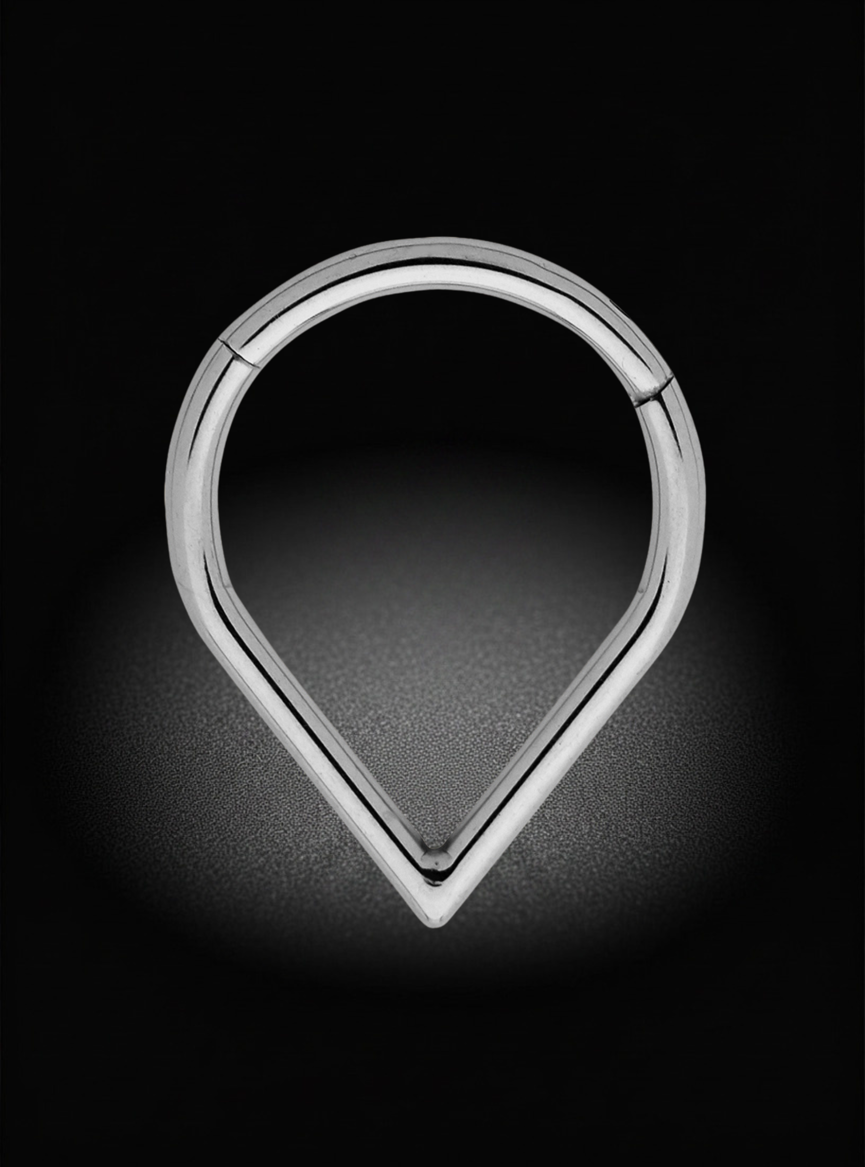 A polished silver-tone metal ring with a teardrop or map-pin shape, shown on a dark black background with a soft spotlight effect.