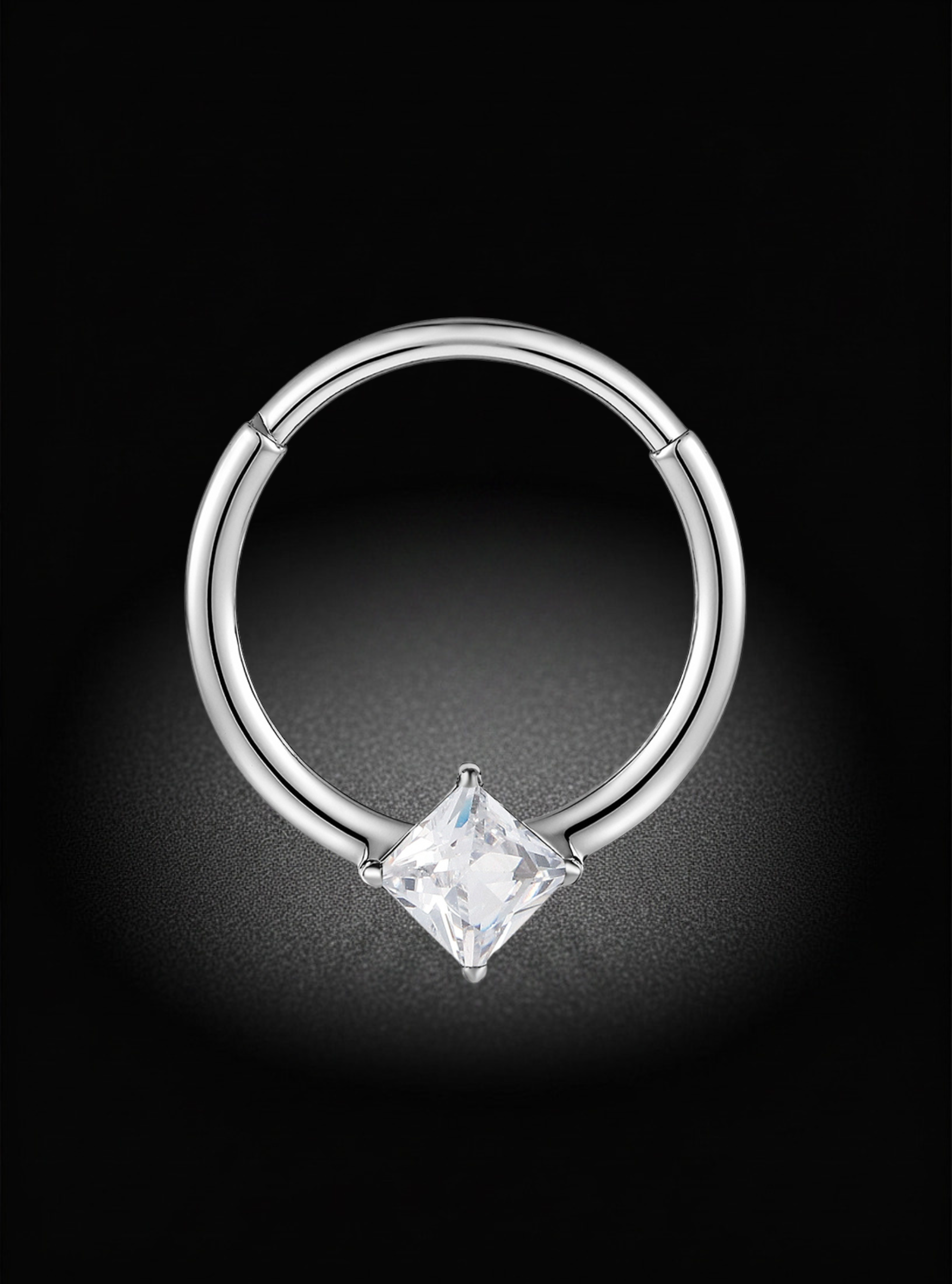 A silver-toned hoop piercing ring with a princess-cut clear gemstone, shown against a dark black background with a soft spotlight effect.
